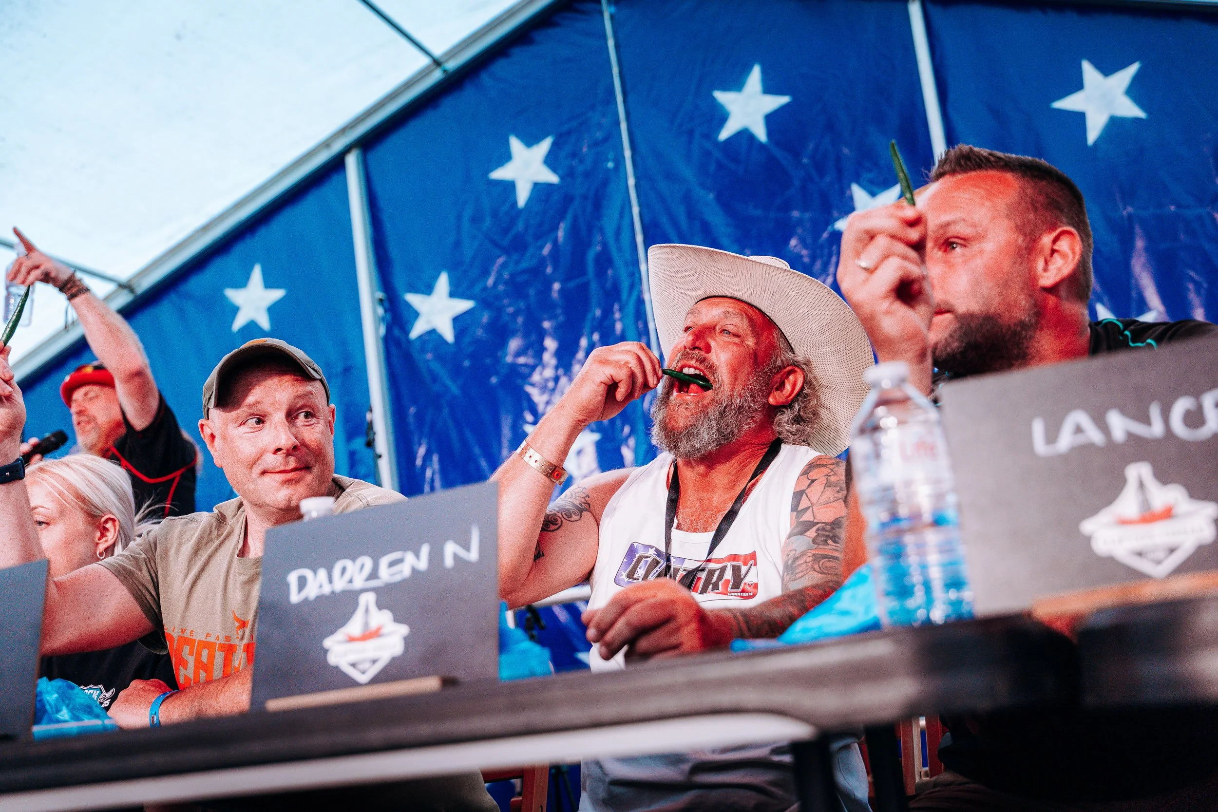 Contestants sample intense peppers at the long table during a live chilli eating competition hosted by the Clifton Chilli Club at Rock n Ribs. High-energy food entertainment and summer festival stage photography by Will Bailey.