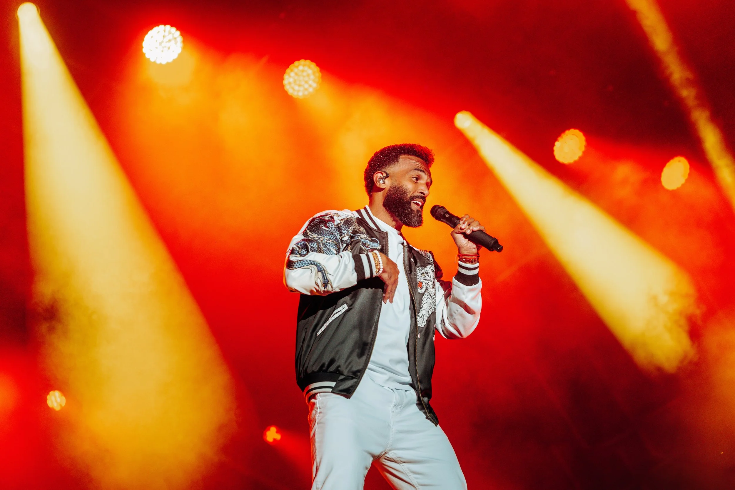 R&B artist Craig David energizing the crowd from the main stage under vibrant orange concert lighting at CarFest in Basingstoke, Hampshire. High-profile celebrity performance and summer festival stage photography by Will Bailey.