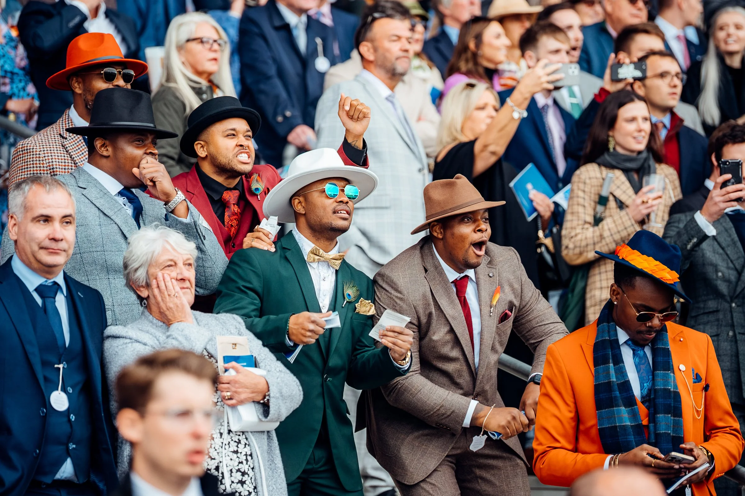 An animated crowd of spectators react with anticipation as the horses sprint past the main grandstand terraces at a Berkshire racing fixture. Candid human emotion and premier public festival photography by Will Bailey.