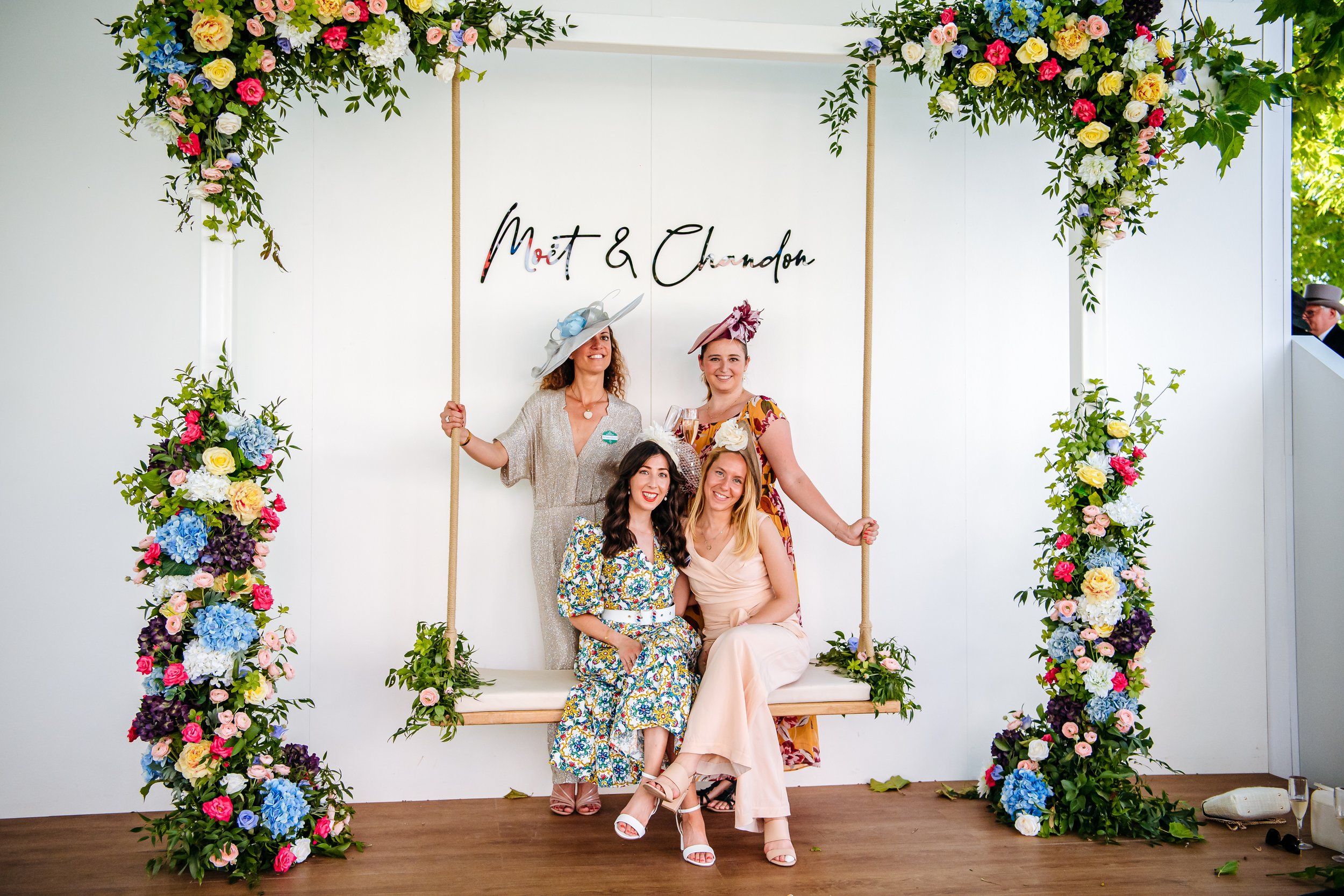 Guests posing at the custom-built floral swing photo activation inside the Moët & Chandon sponsor enclosure at Royal Ascot, Berkshire. Luxury brand experience and experiential marketing activation photography by Will Bailey.