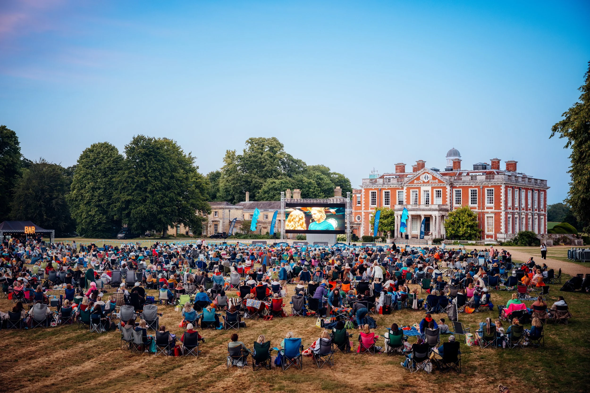 An expansive crowd gathered on the manicured lawns of the historic Stansted Park mansion house for a sunset open-air cinema festival event. Heritage venue programming and regional public lifestyle photography by Will Bailey.
