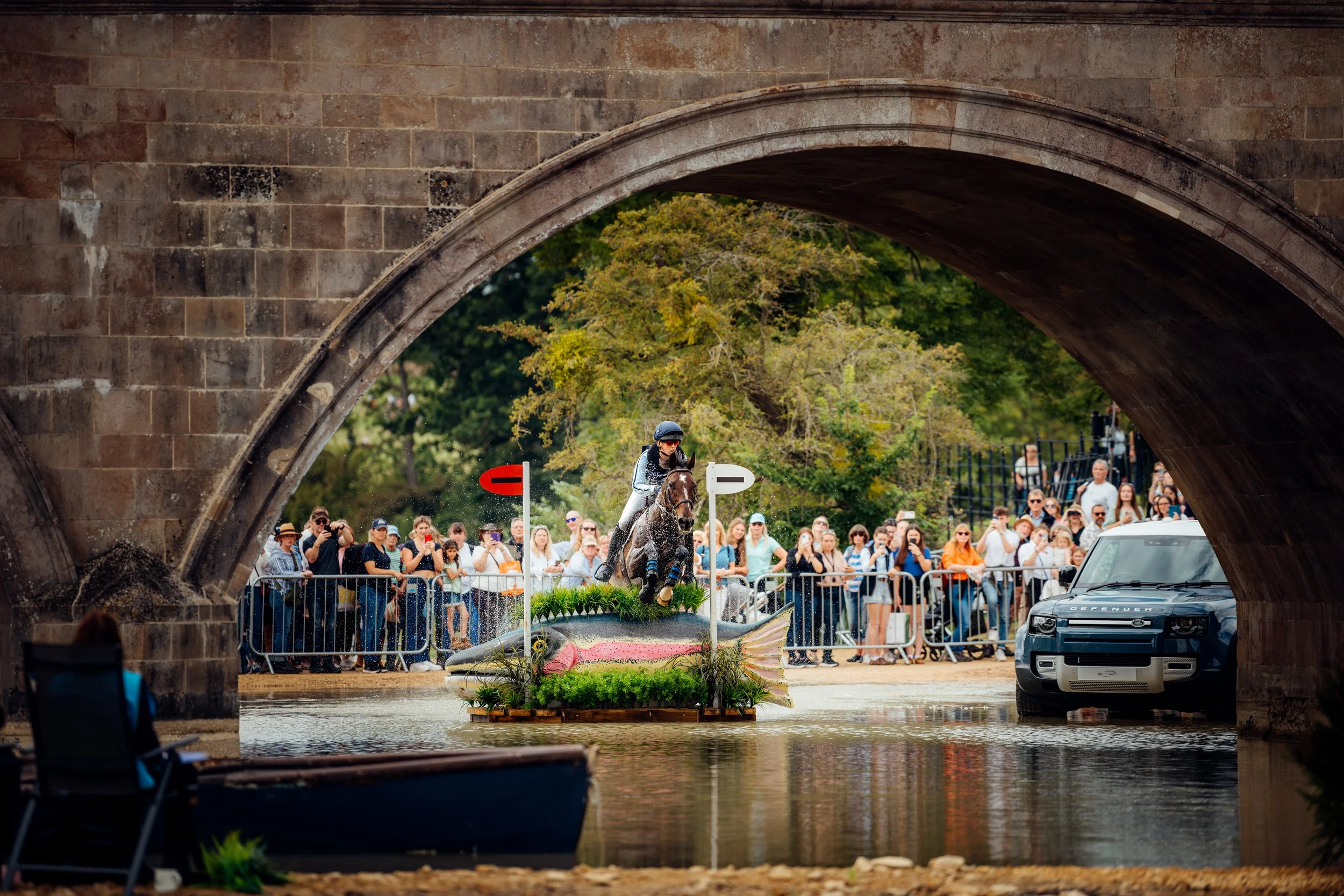 An equestrian competitor clearing a custom fish-shaped water jump obstacle beneath an historic stone archway during the cross-country phase of a prestigious horse trials event. Elite country sports and estate eventing photography by Will Bailey.