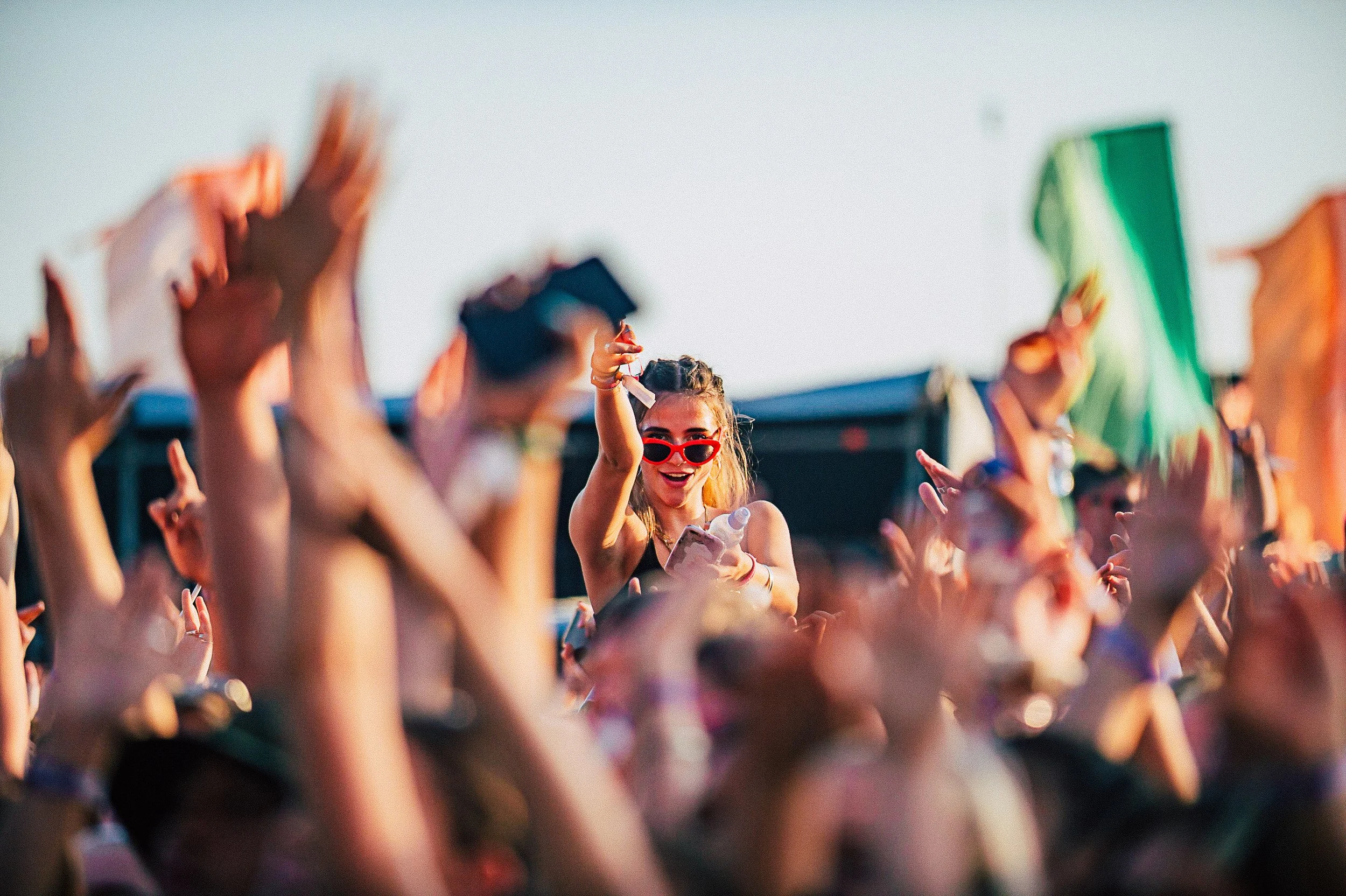 Selective-focus festival crowd portrait of a girl wearing red sunglasses waving among cheering hands at a summer music festival, captured by Will Bailey.