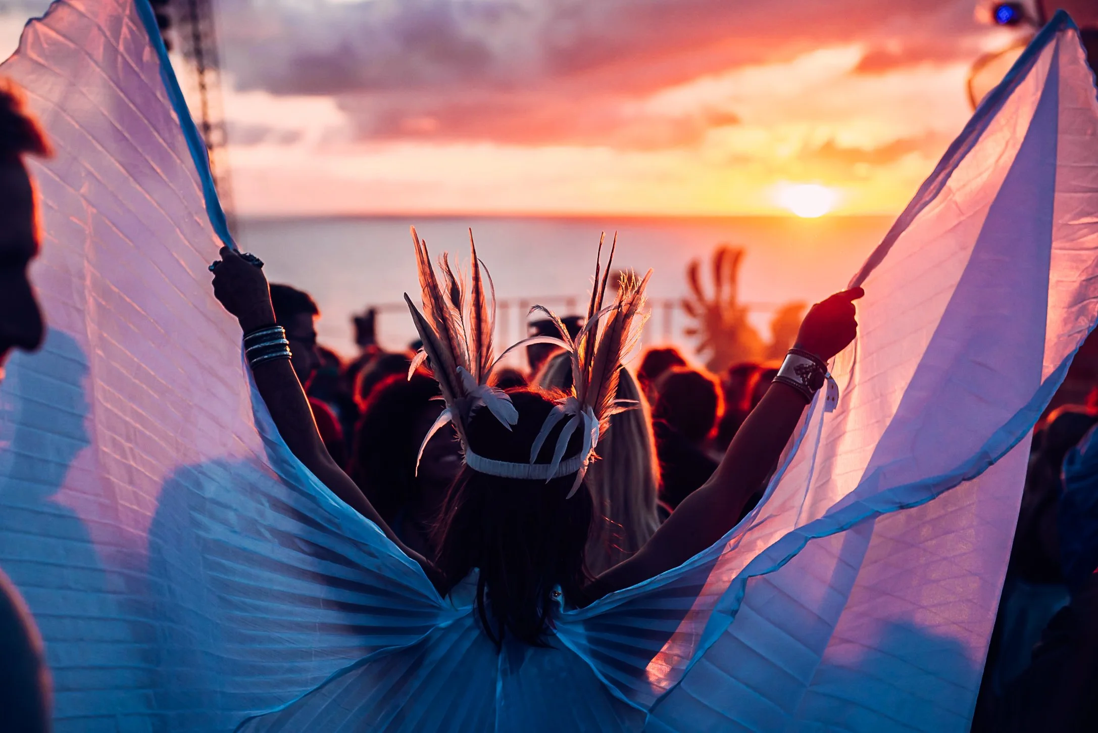 Atmospheric sunset festival lifestyle photography of a dancer wearing a feather headpiece and silk wings at Boardmasters Festival in Cornwall, captured by Will Bailey.