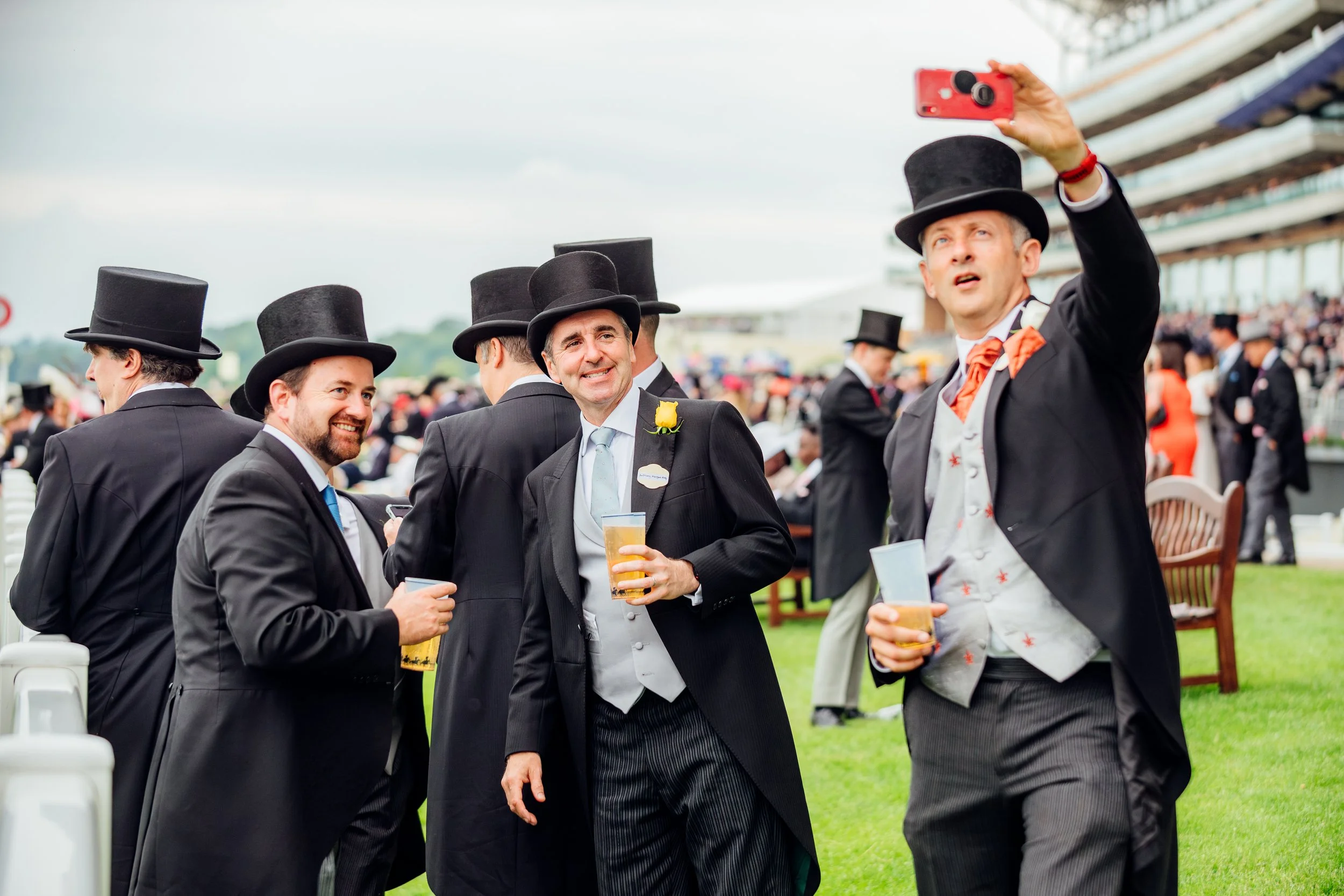 Racegoers in tailored morning dress and silk top hats capturing a candid group selfie by the rails at Ascot Racecourse, Berkshire. High-end social lifestyle and summer seasonal tradition photography by Will Bailey.