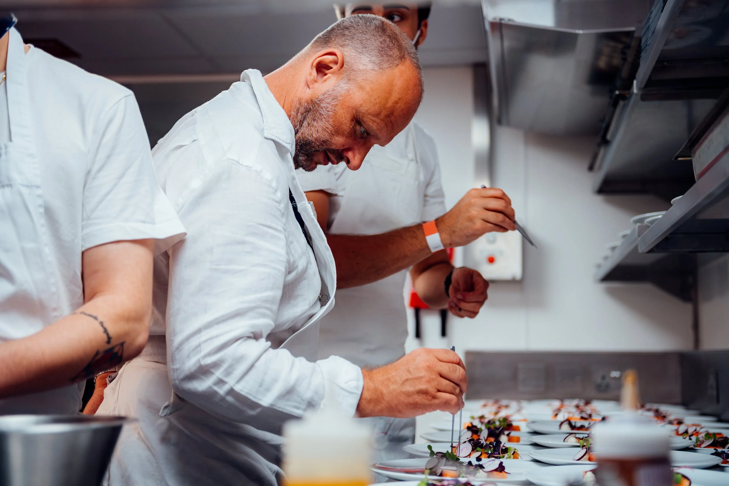 A professional culinary team meticulously plating fine-dining dishes behind the scenes in a high-volume hospitality kitchen at Royal Ascot. Fine-dining lifestyle and luxury event catering photography by Will Bailey.