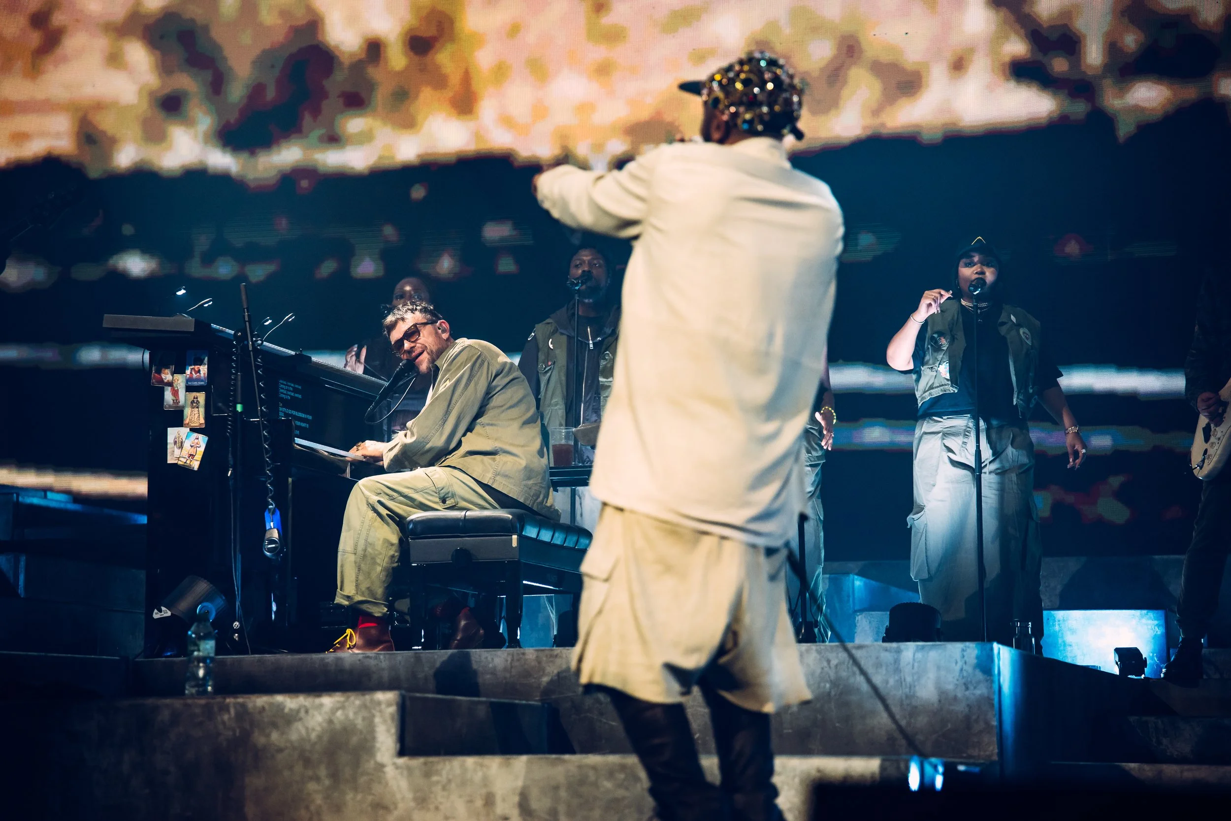Damon Albarn sitting at the piano and singing live while performing on stage with backing vocalists at the Utilita Arena Cardiff. Comprehensive arena tour documentary and editorial live music performance photography by Will Bailey.