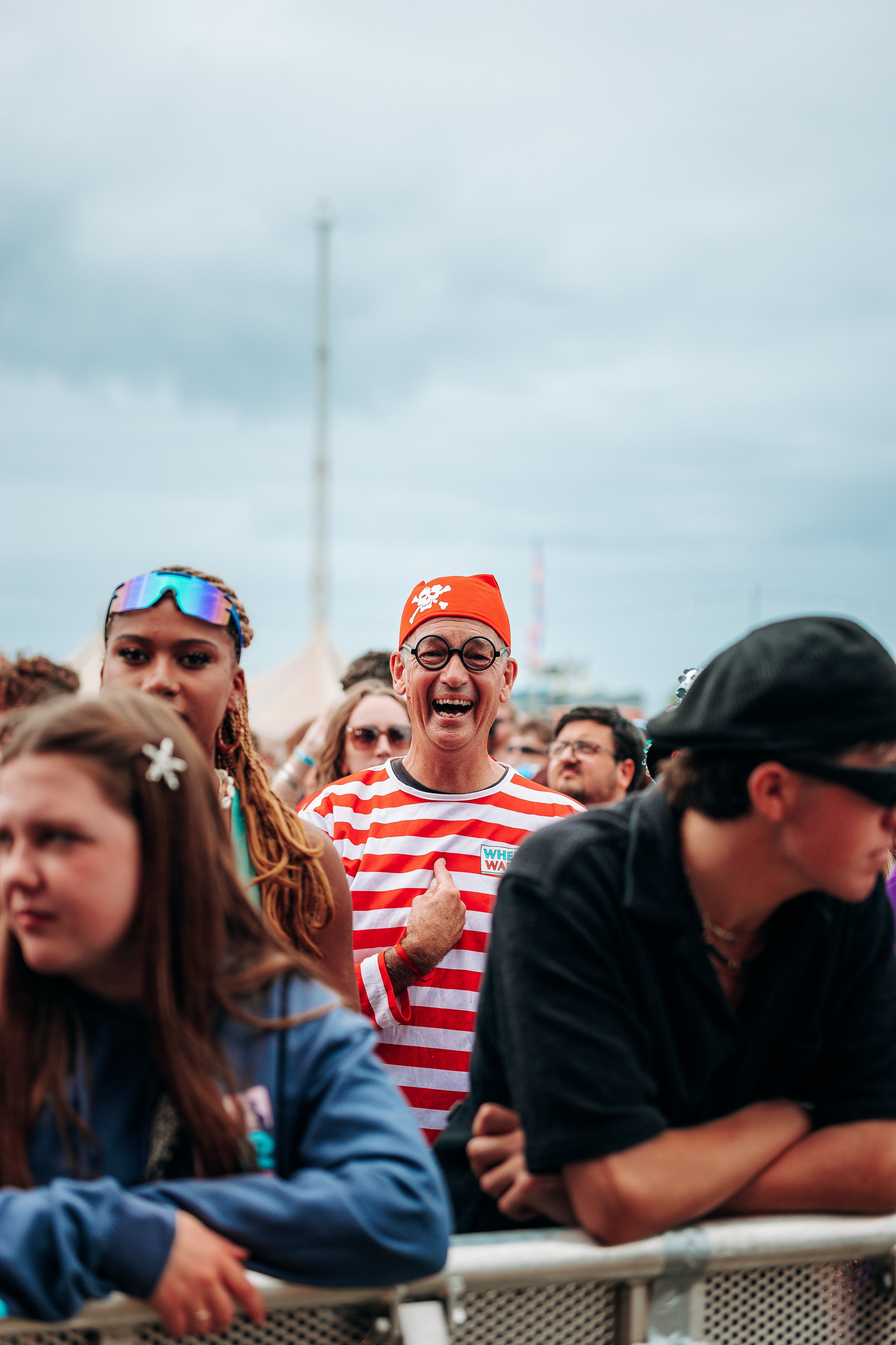 A music fan dressed up in a classic "Where’s Wally?" costume smiling at the front barrier during an outdoor summer festival main stage performance. Candid crowd interaction and public event fan culture photography by Will Bailey.