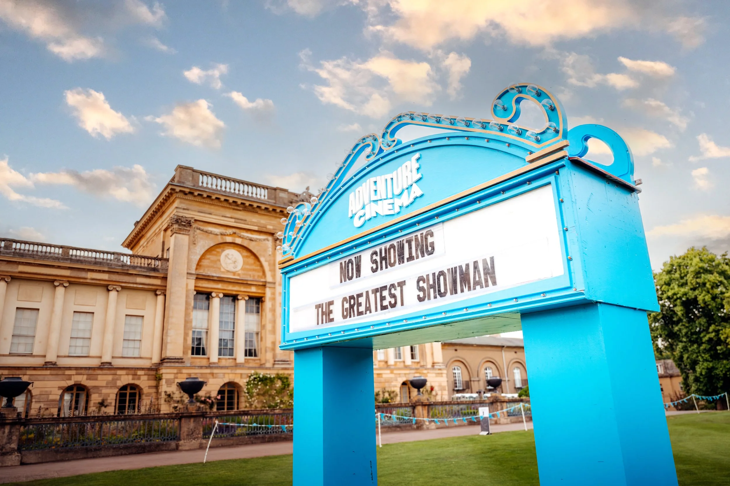 A retro illuminated marquee billboard advertising an Adventure Cinema summer screening of The Greatest Showman, set against the historic facade of Stowe House in Buckinghamshire. Heritage estate programming and outdoor lifestyle cinema photography by