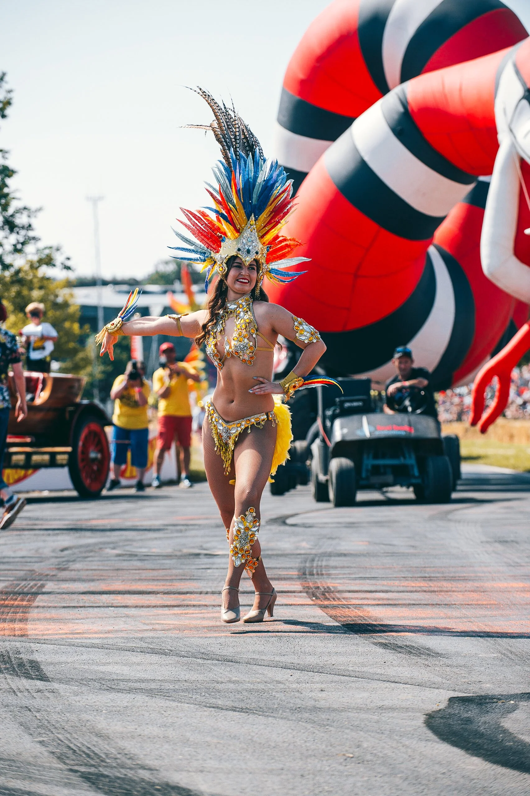 A carnival dancer in an elaborate feathered headdress performing on the track during a public parade activation at CarFest. High-energy festival entertainment and vibrant summer celebration photography by Will Bailey.