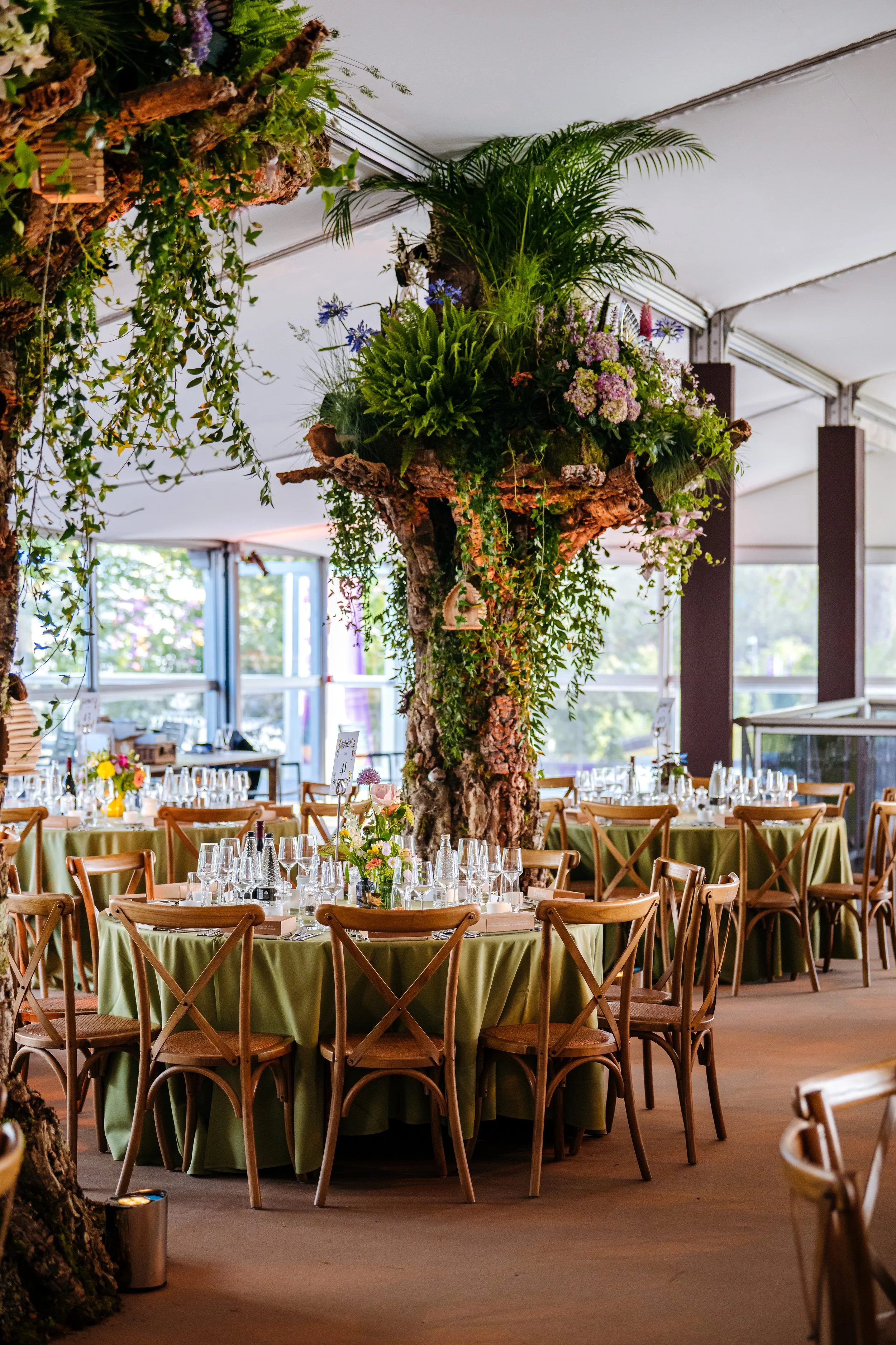 An immaculately designed dining marquee pavilion featuring living tree installations and organic floral centerpieces at the RHS Chelsea Flower Show in London. Luxury event design, high-end hospitality catering, and pavilion interior photography by Wi