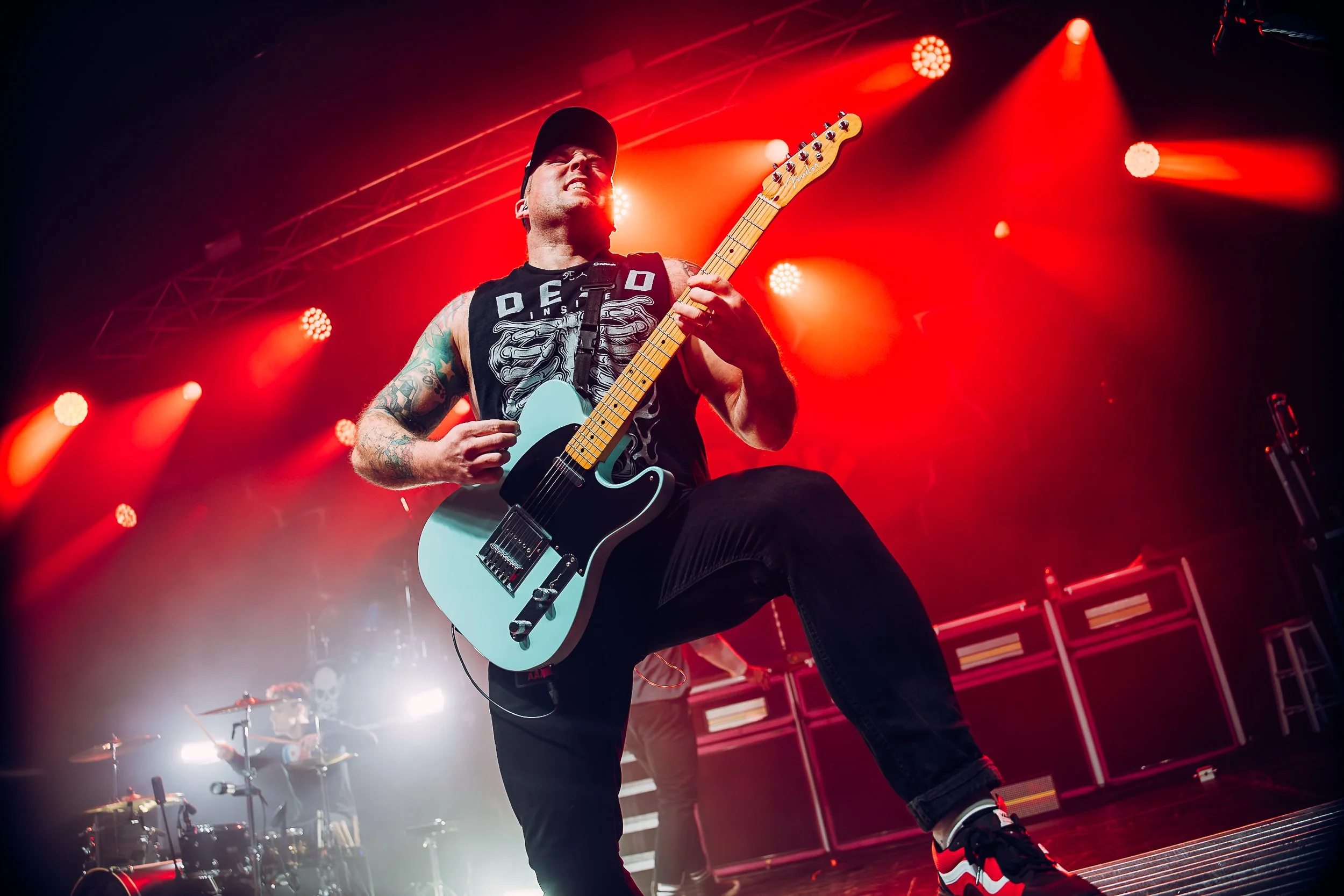 Tom Newton, lead guitarist of pop-punk group The Bottom Line playing a baby blue electric guitar under vibrant red stage wash at the Manchester Academy music venue. Professional national tour coverage and live band promotion by official tour photogra