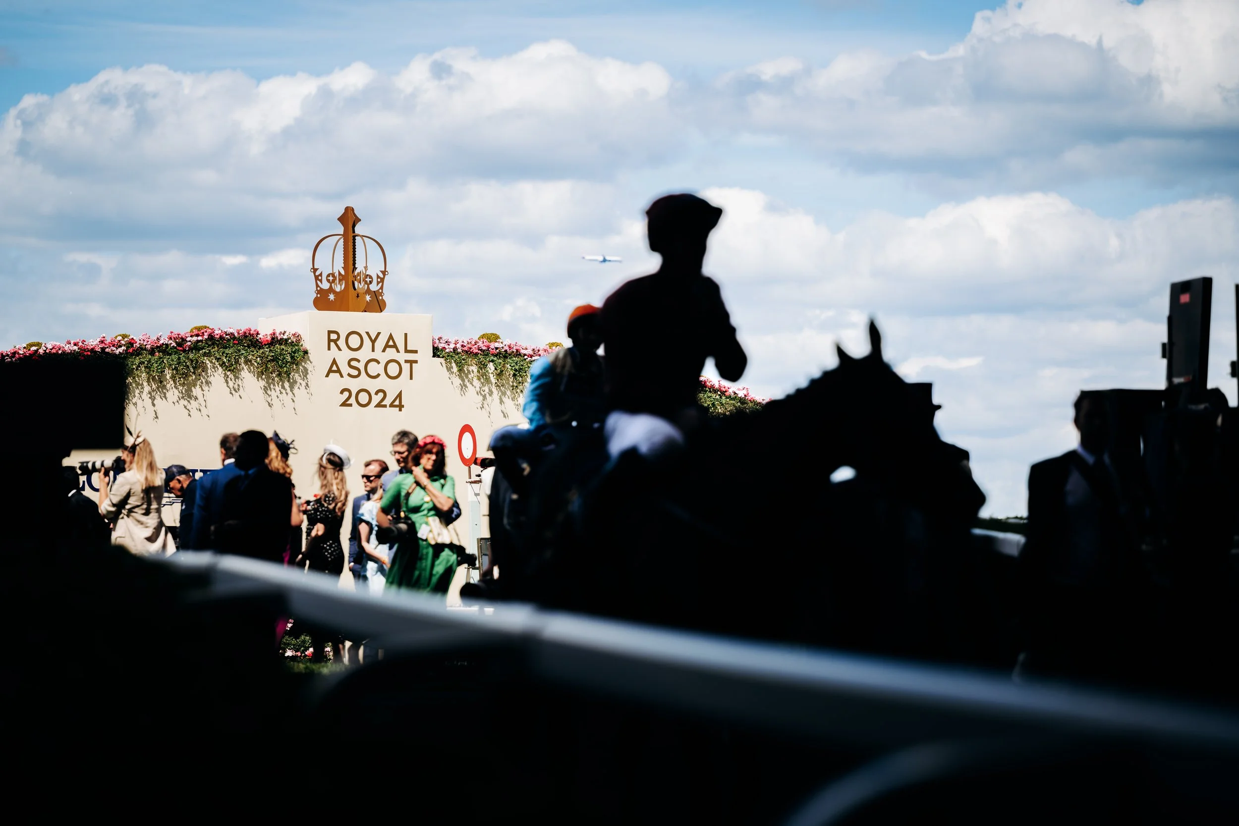A silhouette of horses and jockeys moving past the gilded venue signage against a dramatic sky at Ascot Racecourse, Berkshire. Fine-art sports documentary and professional equestrian event photography by Will Bailey.