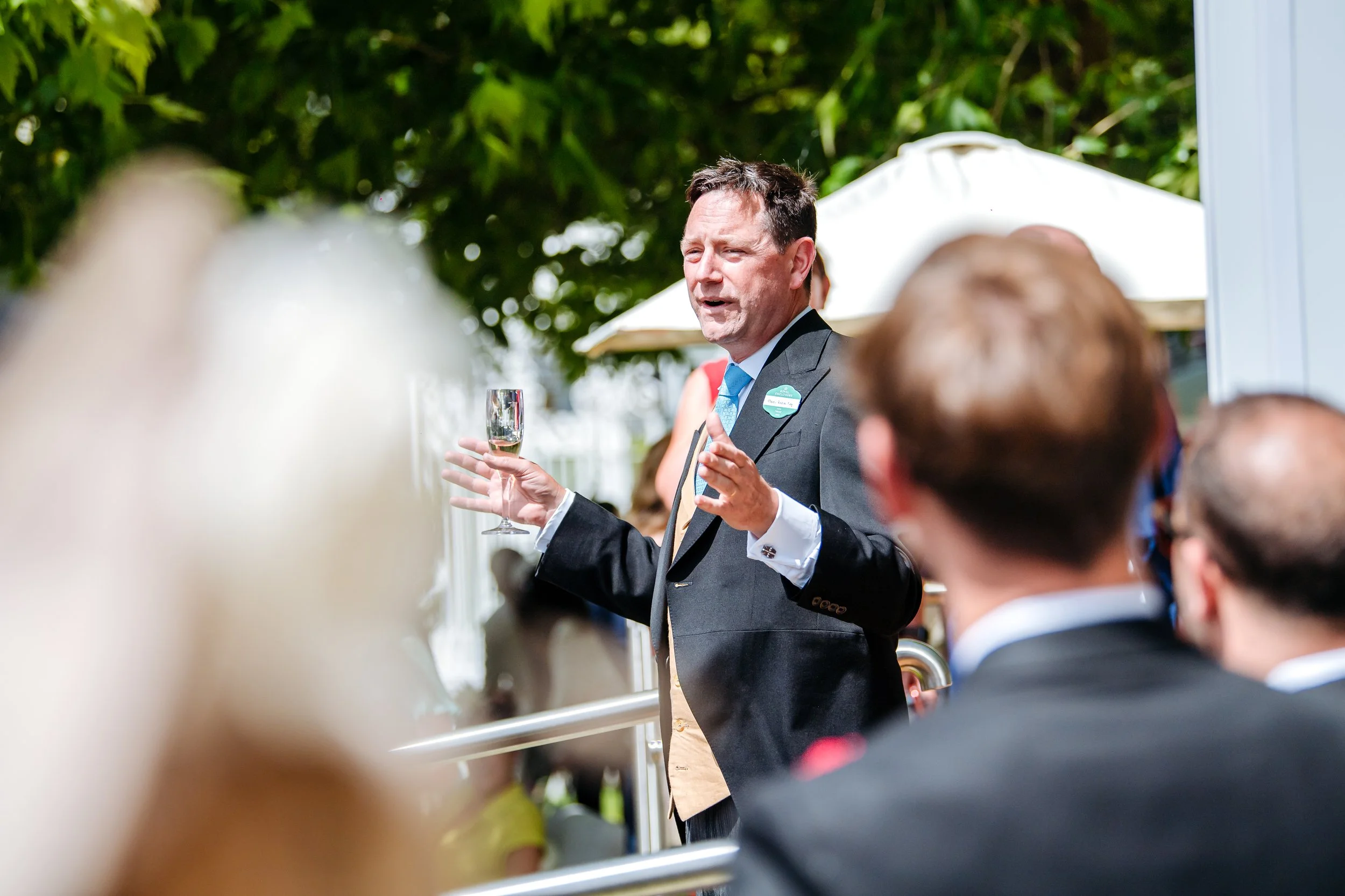 A guest delivering an eloquent toast with a champagne flute during a private hospitality reception at Royal Ascot in Berkshire. Premium summer season social calendar and luxury lifestyle event photography by Will Bailey.