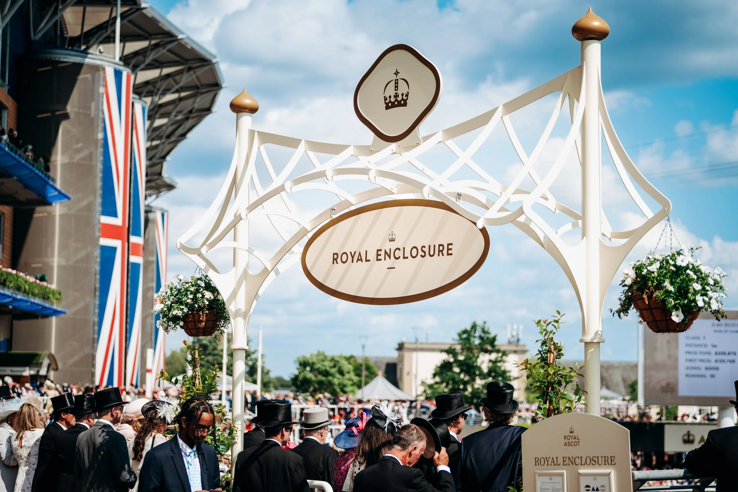Spectators in morning dress passing beneath the cream-colored wrought-iron gates marking the Royal Enclosure entrance at Royal Ascot, Berkshire. British high-society cultural tradition and elite venue lifestyle photography by Will Bailey.