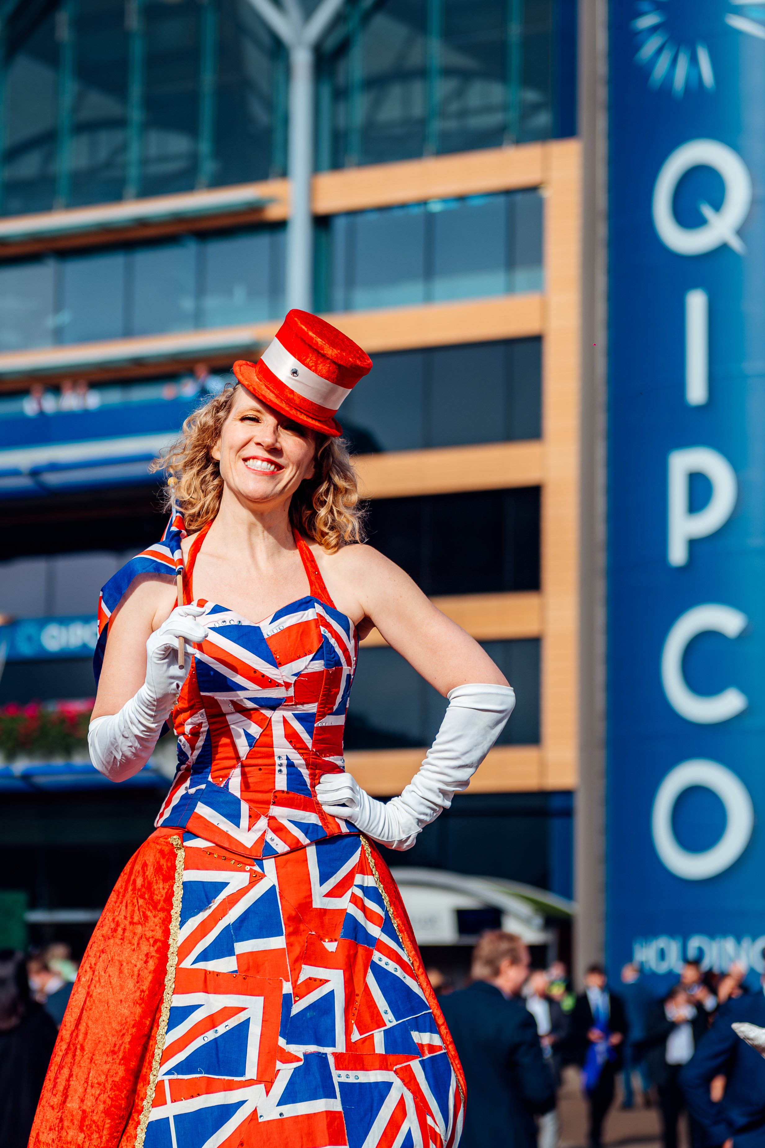 A stilt walker in a glittering Union Jack gown performing for crowds at the QIPCO British Champions Day at Ascot Racecourse, Berkshire. Experiential street theatre and autumn festival entertainment photography by Will Bailey.
