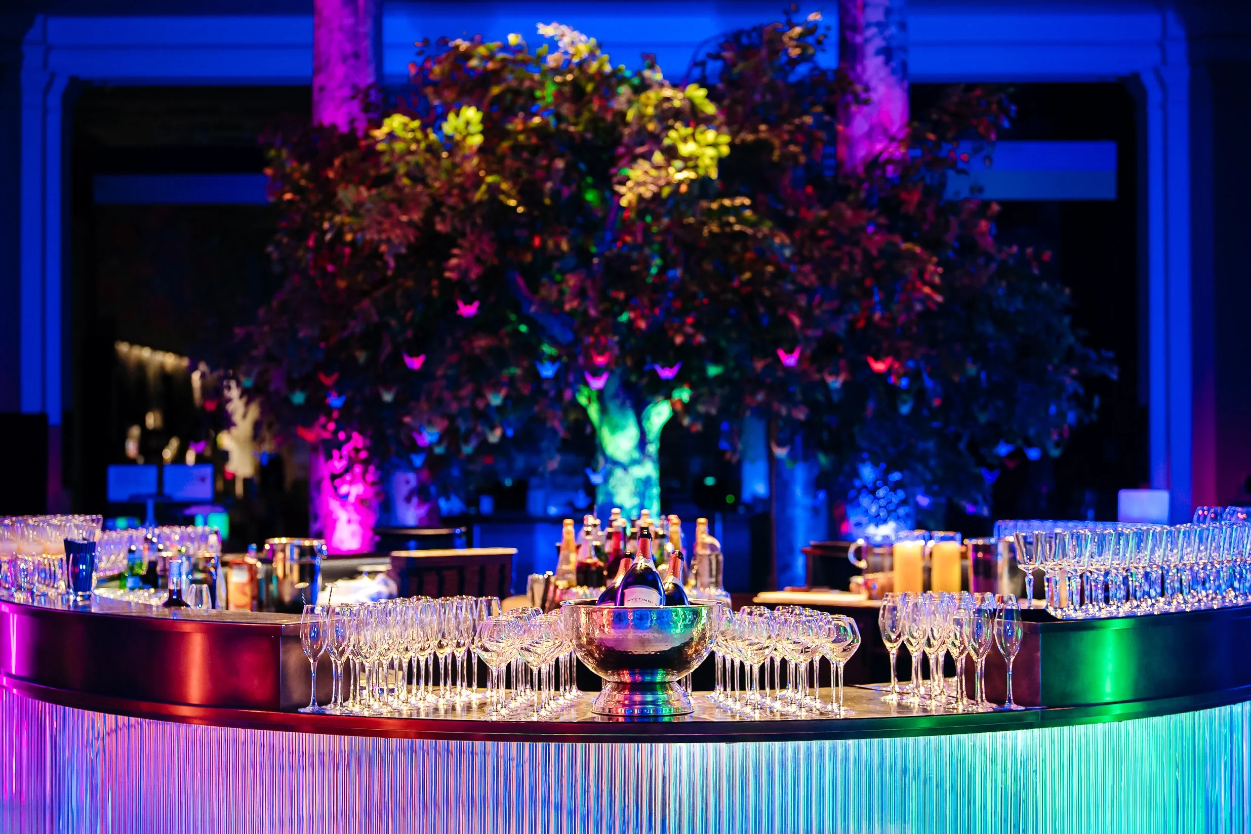 A pristine view of the illuminated circular champagne bar under dramatic event lighting at the Victoria and Albert Museum (V&A) for the Macmillan fundraiser. Premium commercial corporate event photography by Will Bailey.