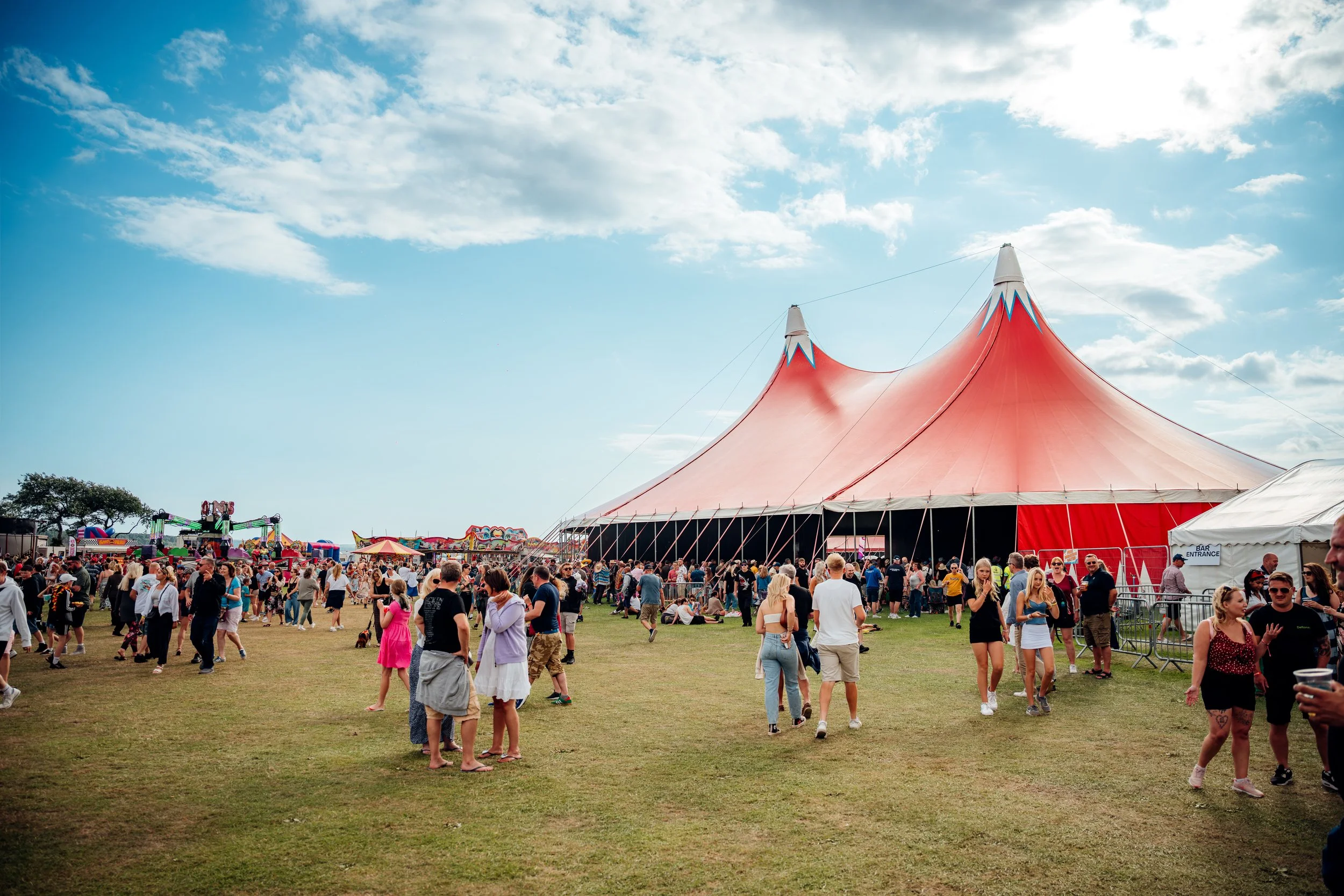 Crowds of families and music fans walking the outdoor fairground and main stage field at the Poole Harbour Festival. Dynamic summer music festival and regional public event photography by Will Bailey.
