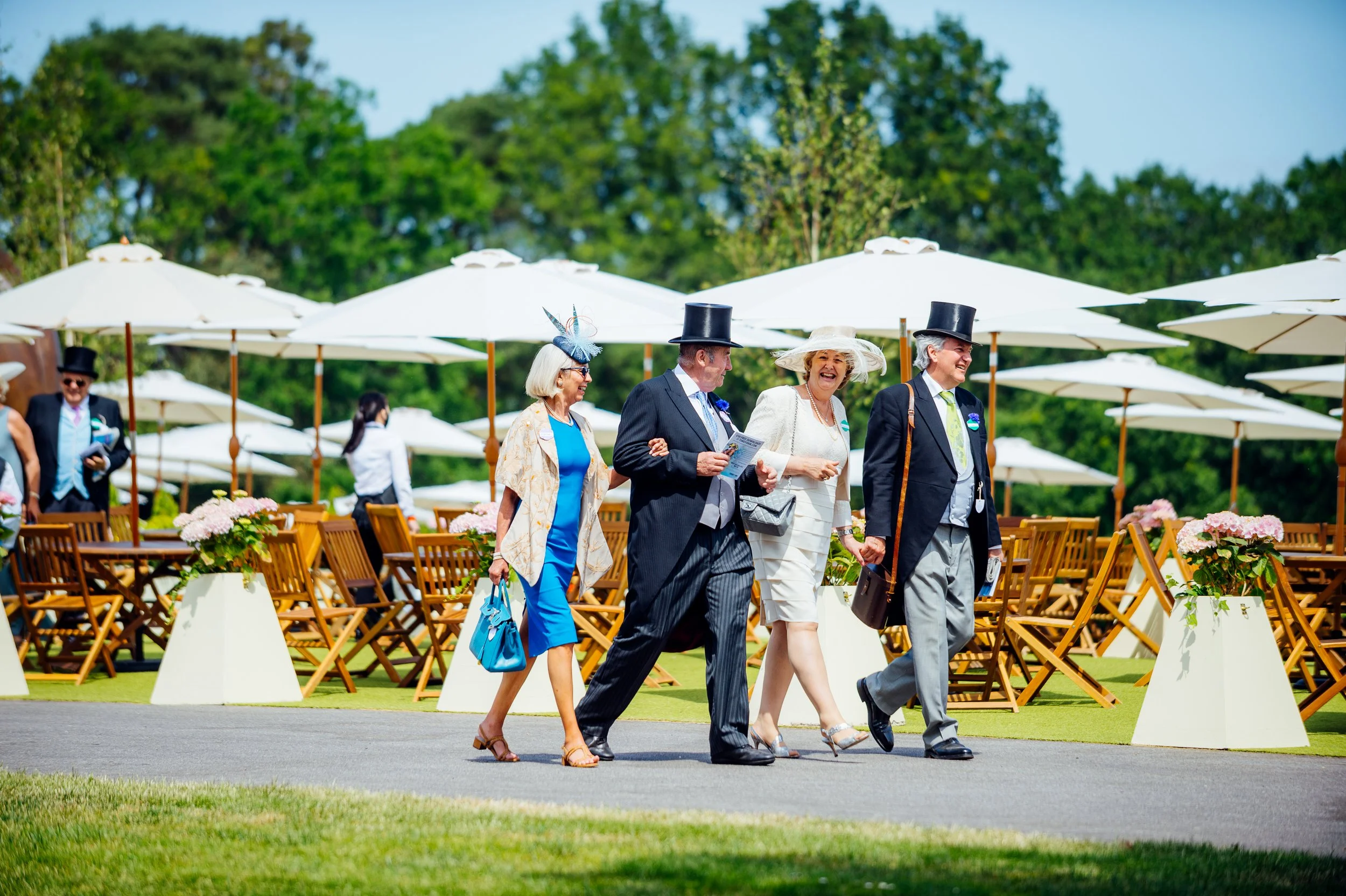 Racegoers in elegant formalwear and top hats walking past luxury umbrella seating in the Royal Enclosure at Royal Ascot. British high-society sporting season and luxury lifestyle event photography by Will Bailey.
