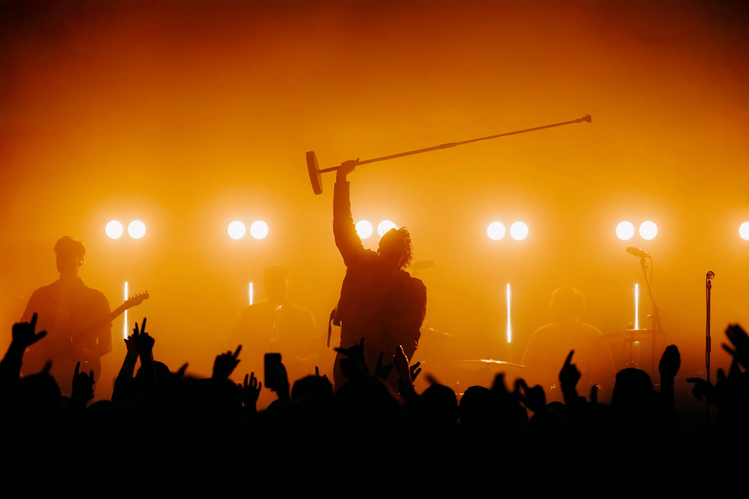A dramatic orange silhouette of Kids In Glass Houses raising a microphone stand high above a hyped crowd at an independent concert venue in Southampton, Hampshire. High-impact rock concert and creative venue stage lighting photography by Will Bailey.