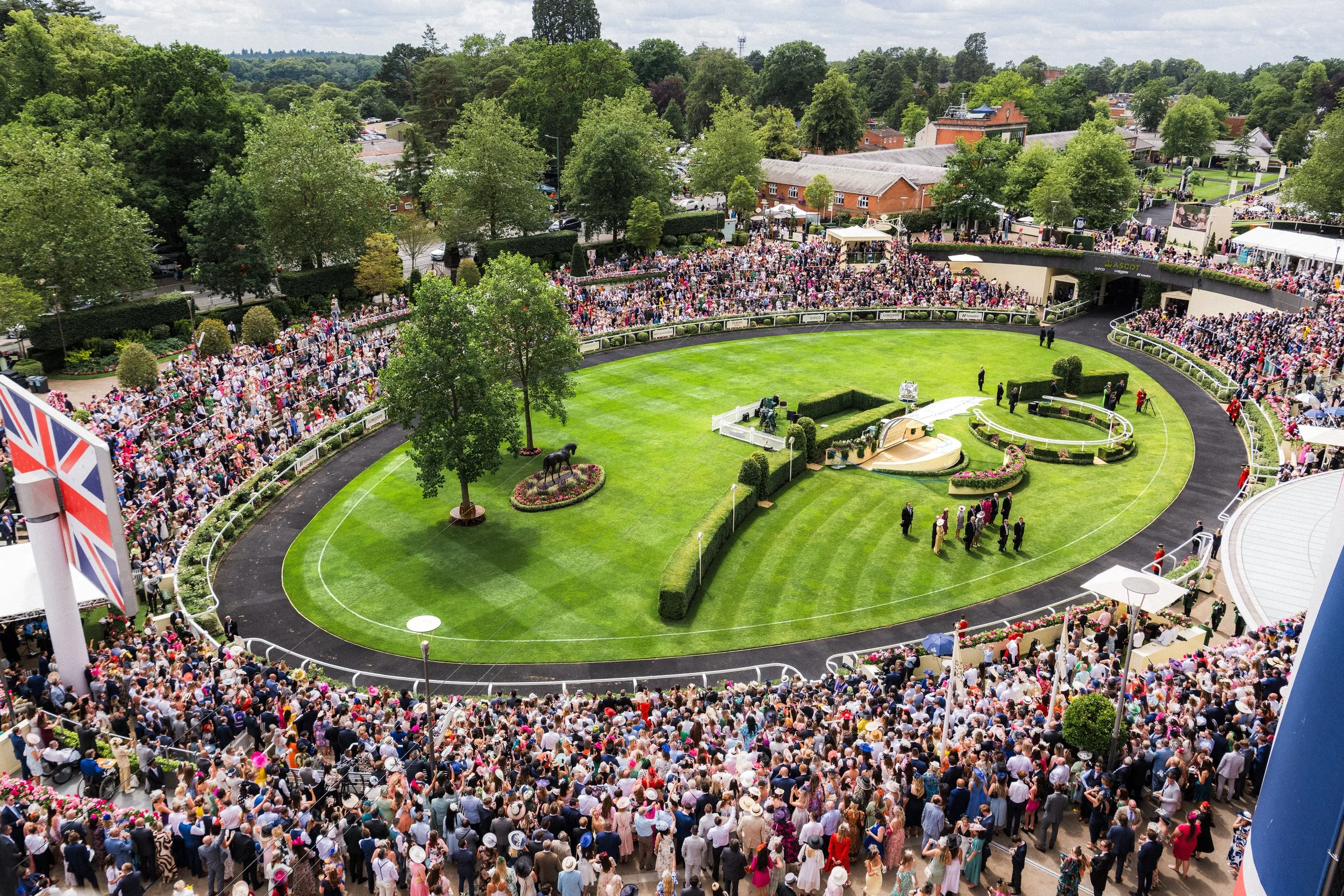 An elevated high-angle view of the packed parade ring and spectator terraces during a major summer race meeting at Ascot, Berkshire. Prestigious venue architecture and large-scale public event photography by Will Bailey.