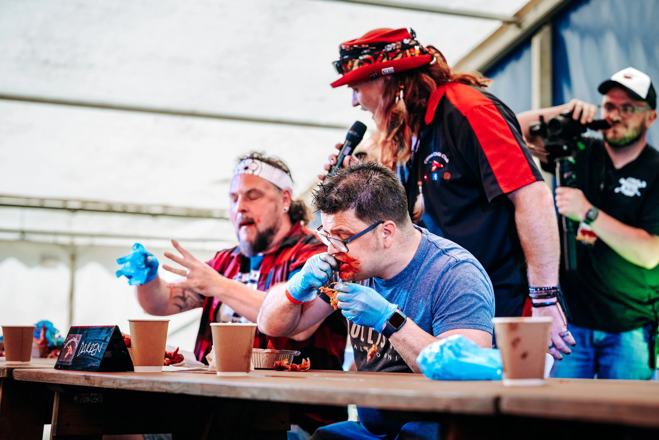 Contestants competing in a high-intensity hot wing eating challenge hosted live by the Clifton Chilli Club on stage at the Rock n Ribs Festival. High-energy public entertainment and food festival action photography by Will Bailey.