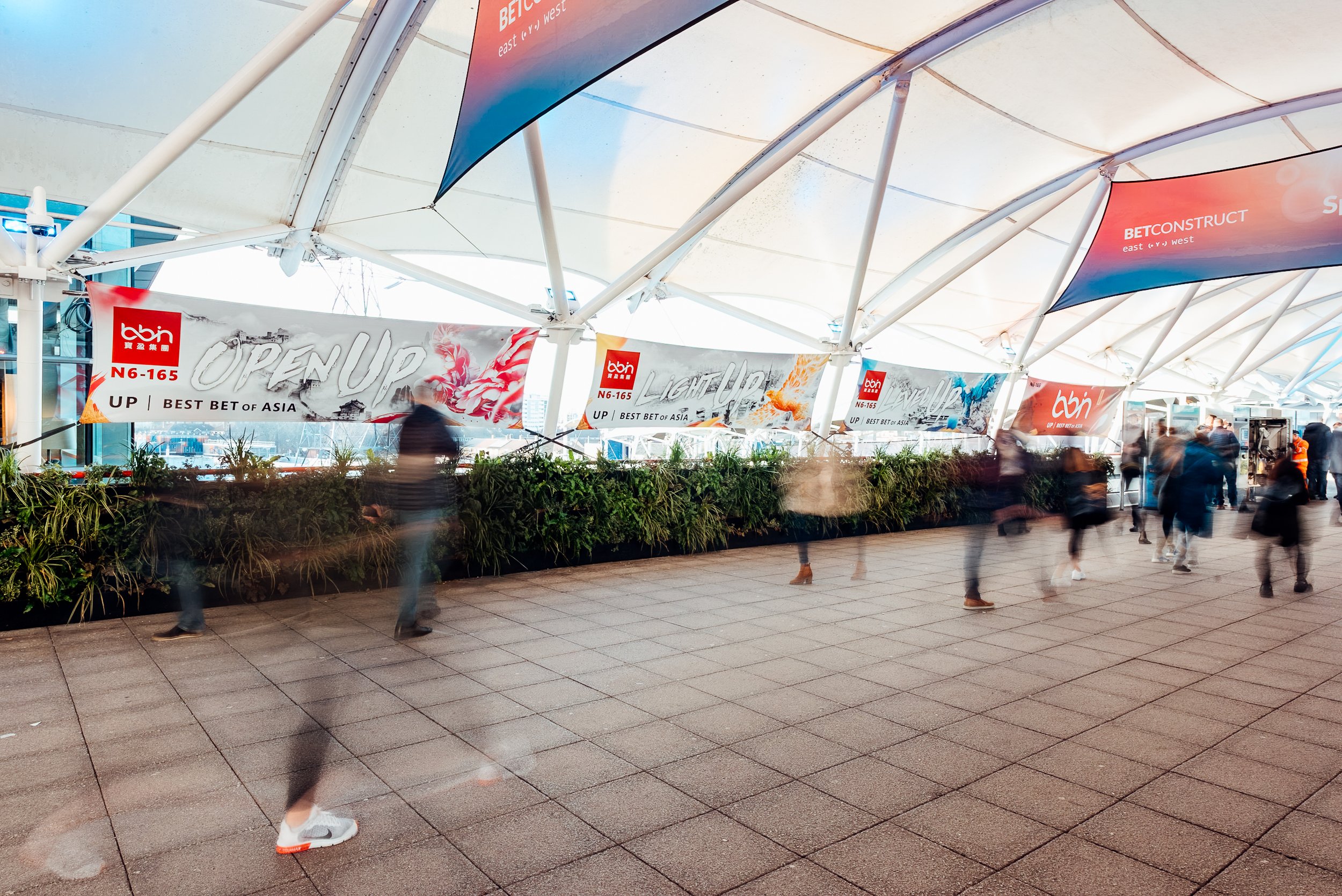 Dynamic attendee motion blur passing the custom BBIN advertising banners at the entrance canopy of ExCeL London during the ICE London expo. Exhibition brand activation photography by Will Bailey.
