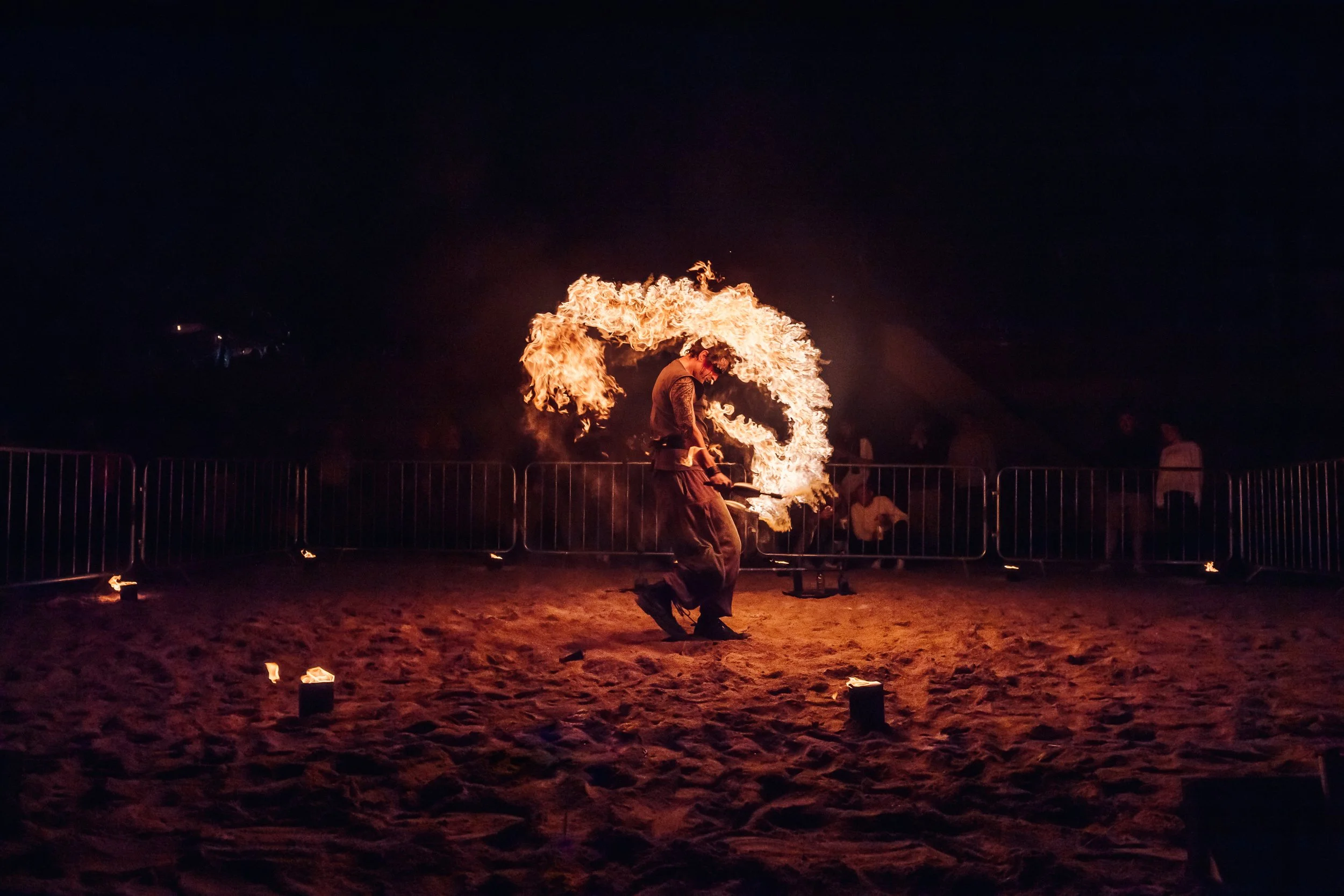 A fire spinner performing an intricate routine on the sand at night during a beachside cultural event in Cornwall. Dramatic nighttime fire performance and coastal festival lifestyle photography by Will Bailey.