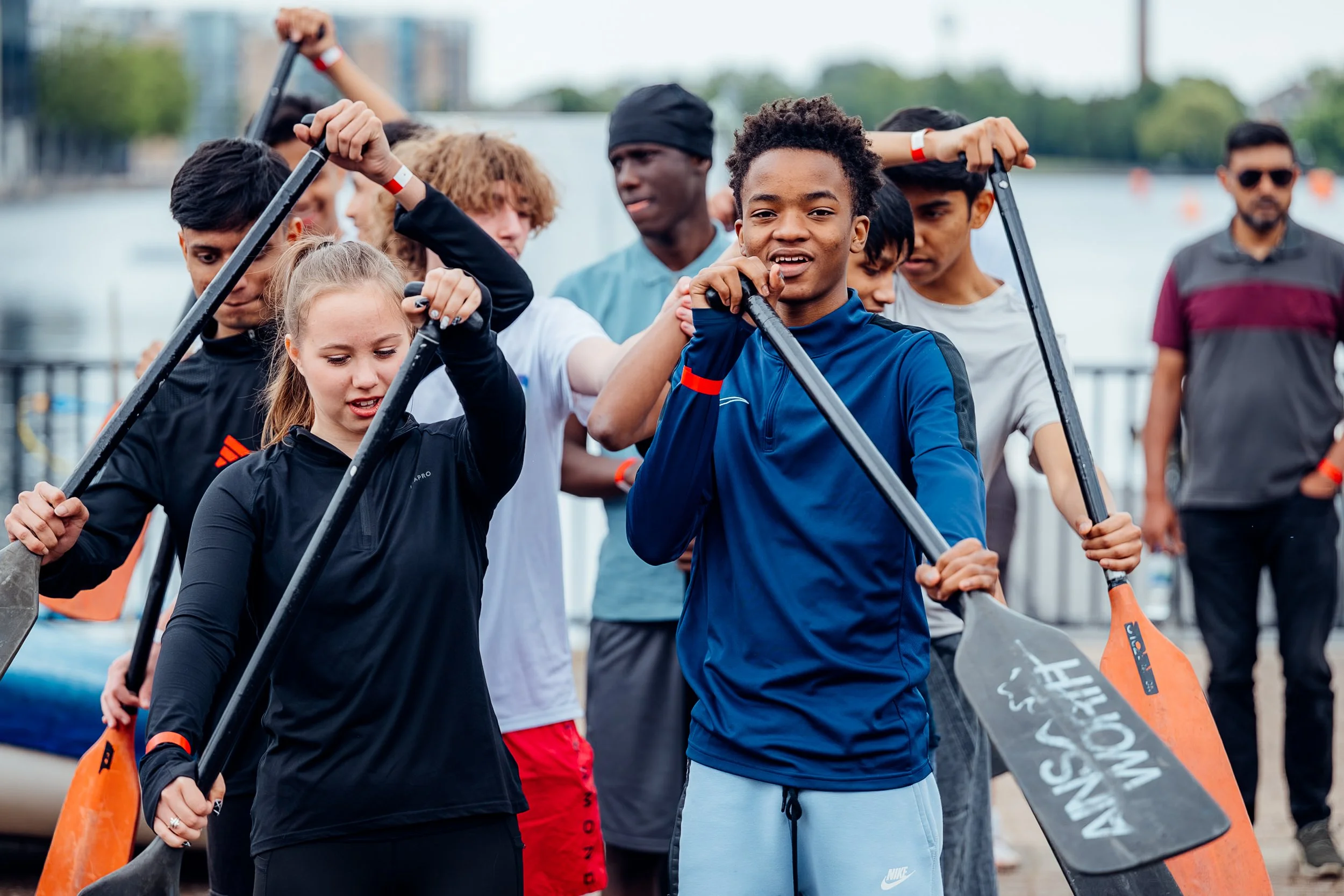 Youth community teams lined up on the dockside with oars, preparing to race at the Far East Consortium (FEC) Charity Dragon Boat Festival in London. CSR and community sports event photography by Will Bailey.