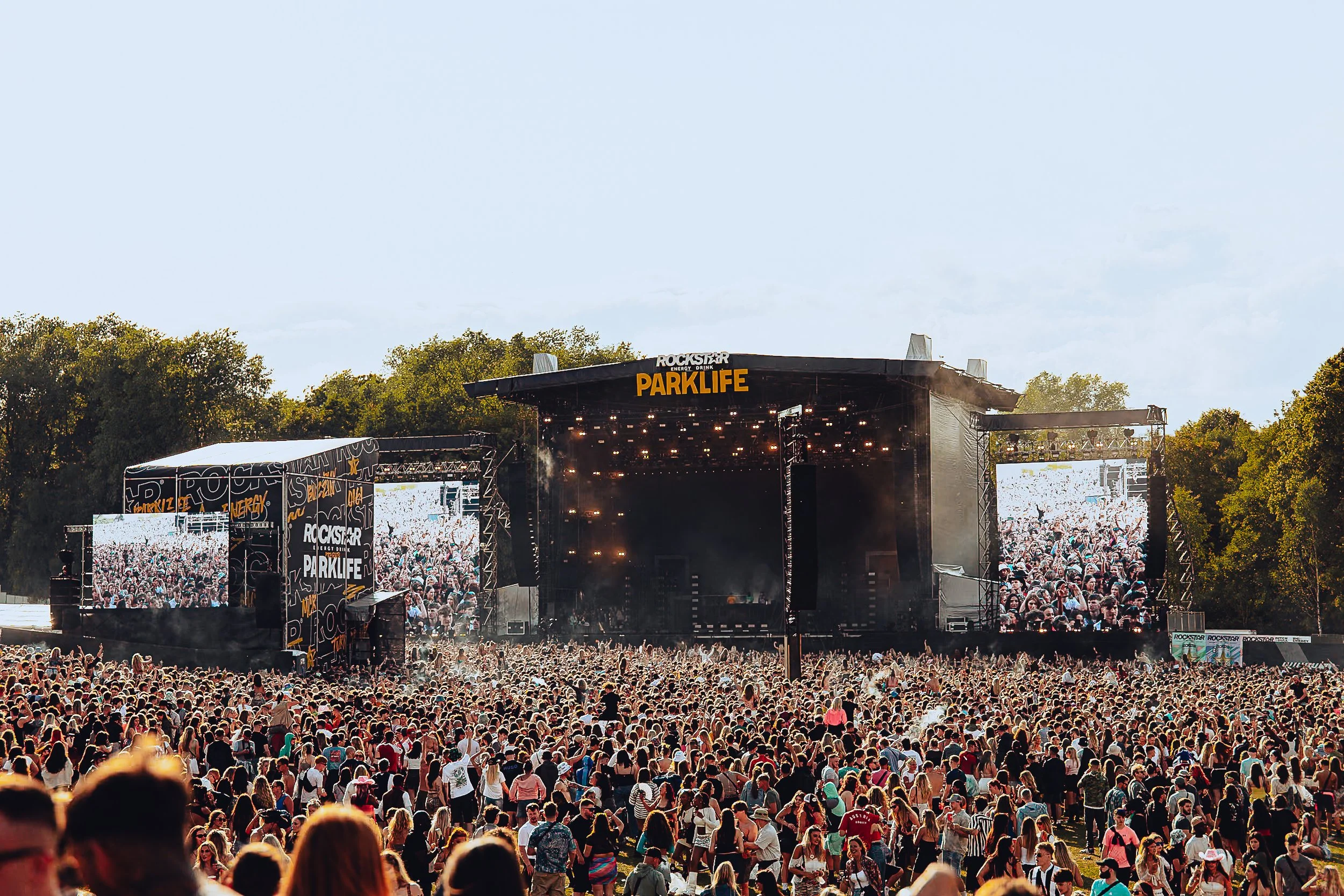 Ultra-wide crowd scale photography of the Rockstar Energy main stage at Parklife Festival in Manchester, capturing thousands of festival attendees in daylight by Will Bailey.