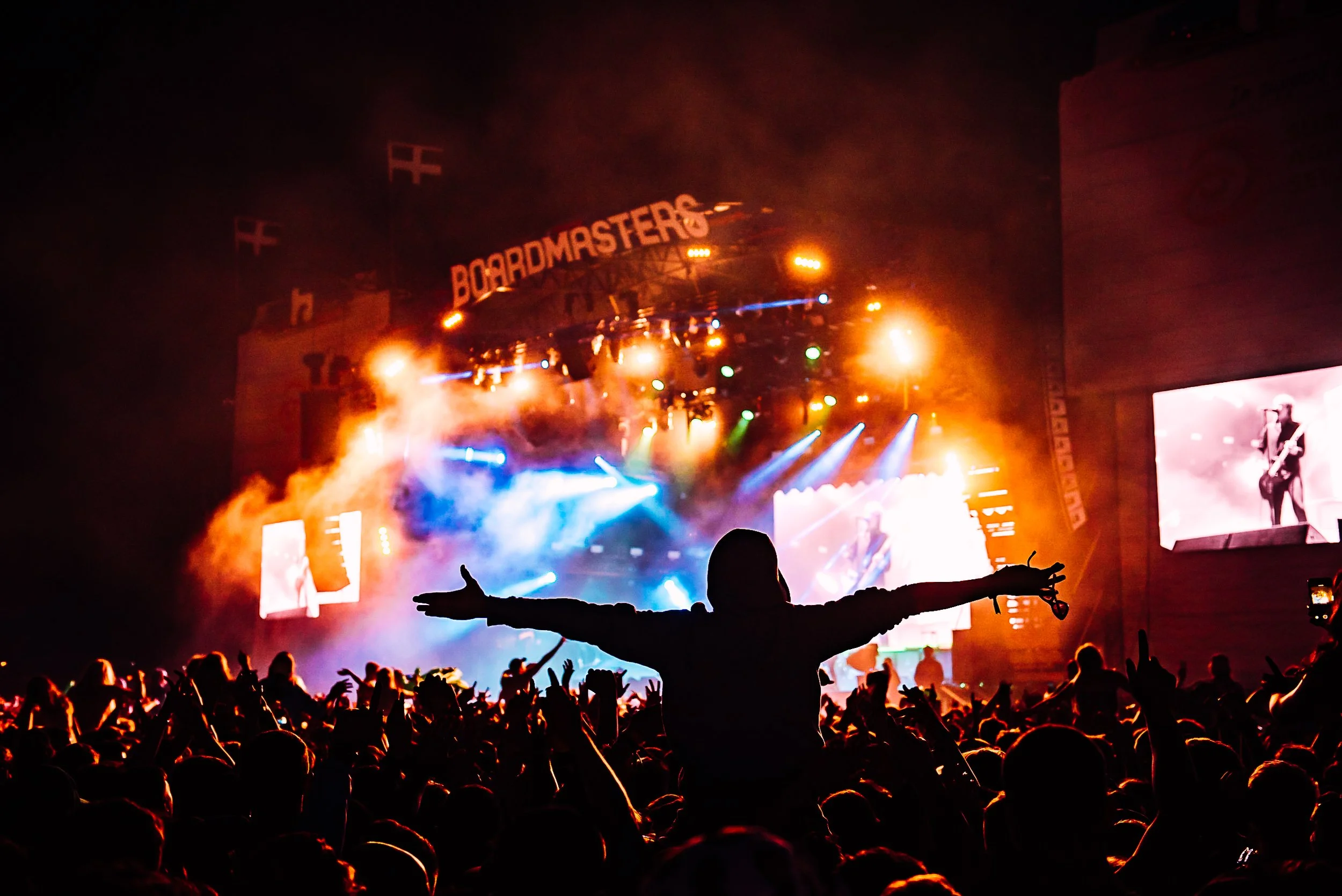 Silhouetted festival attendee cheering on shoulders with arms wide open in front of the illuminated main stage during Catfish and the Bottlemen at Boardmasters, shot by Will Bailey.