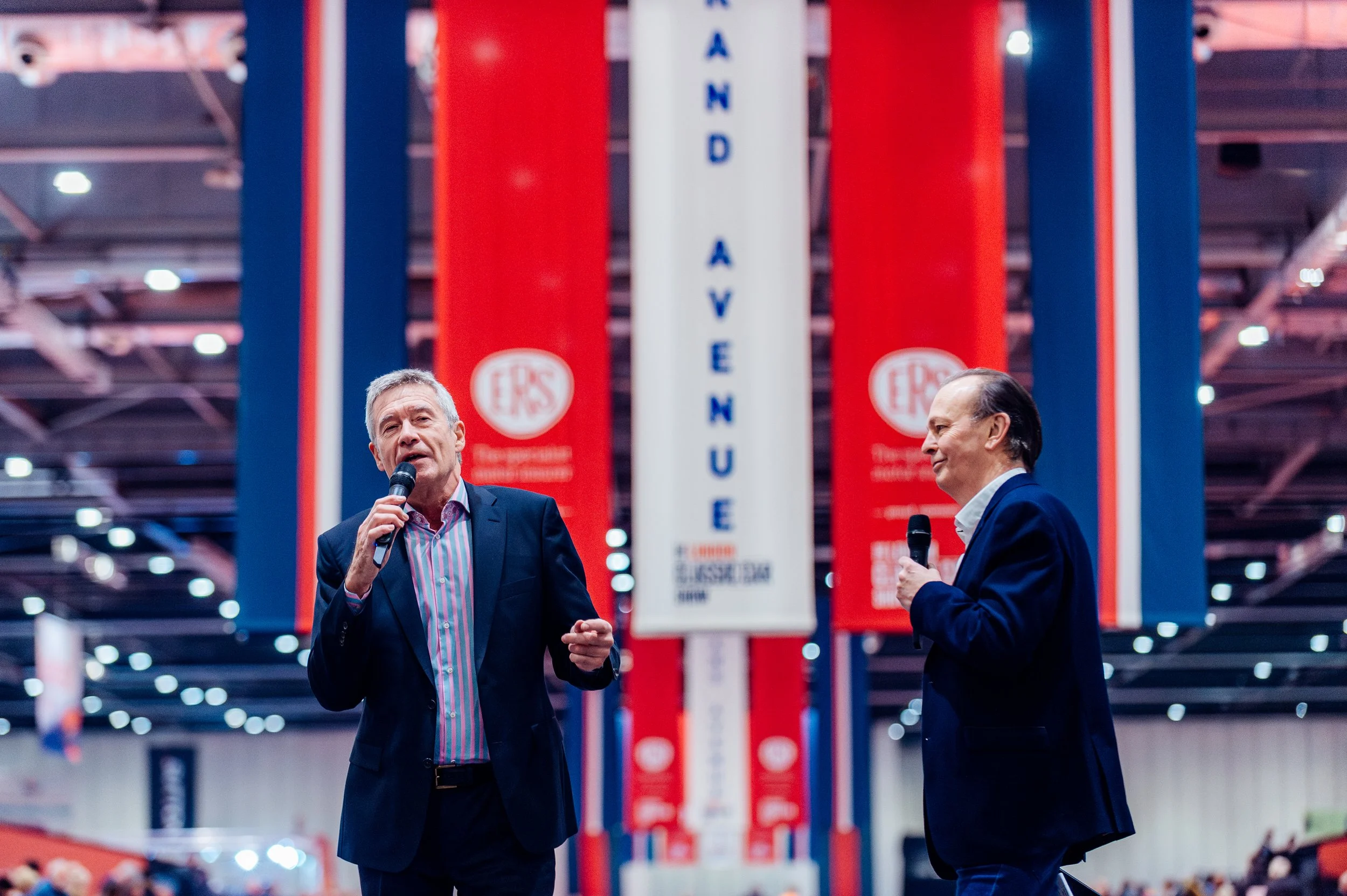 Television presenters Tiff Needell and Quentin Willson speaking live on stage against a classic motorsport backdrop at the London Classic Car Show, ExCeL London. Premium automotive exhibition and event hosting photography by Will Bailey.