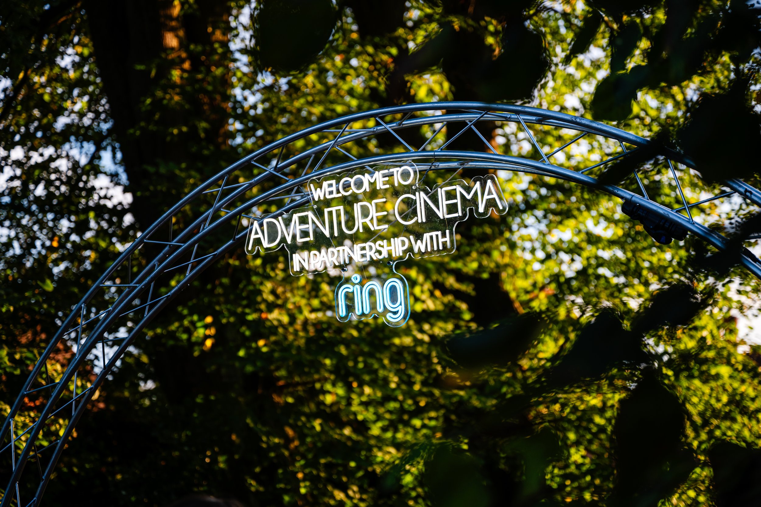 A glowing custom neon sign suspended from an aluminum truss archway marking the public entrance to Adventure Cinema at The Vyne in Basingstoke, Hampshire. Premium experiential event marketing and national trust venue photography by Will Bailey.