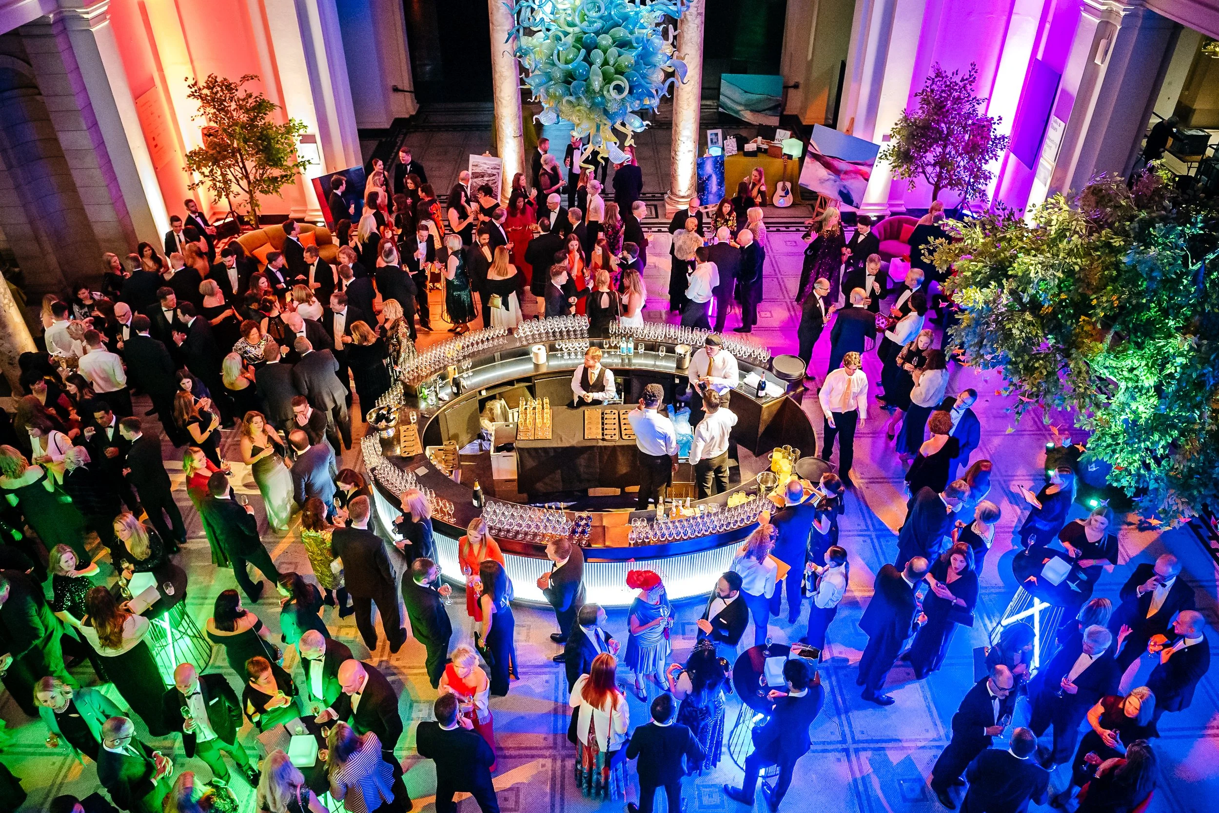 An elevated high-angle view of the bustling black-tie drinks reception surrounding the central cocktail bar during the Macmillan Cancer Support fundraiser at the Victoria and Albert Museum (V&A), London. Luxury gala event photography by Will Bailey.
