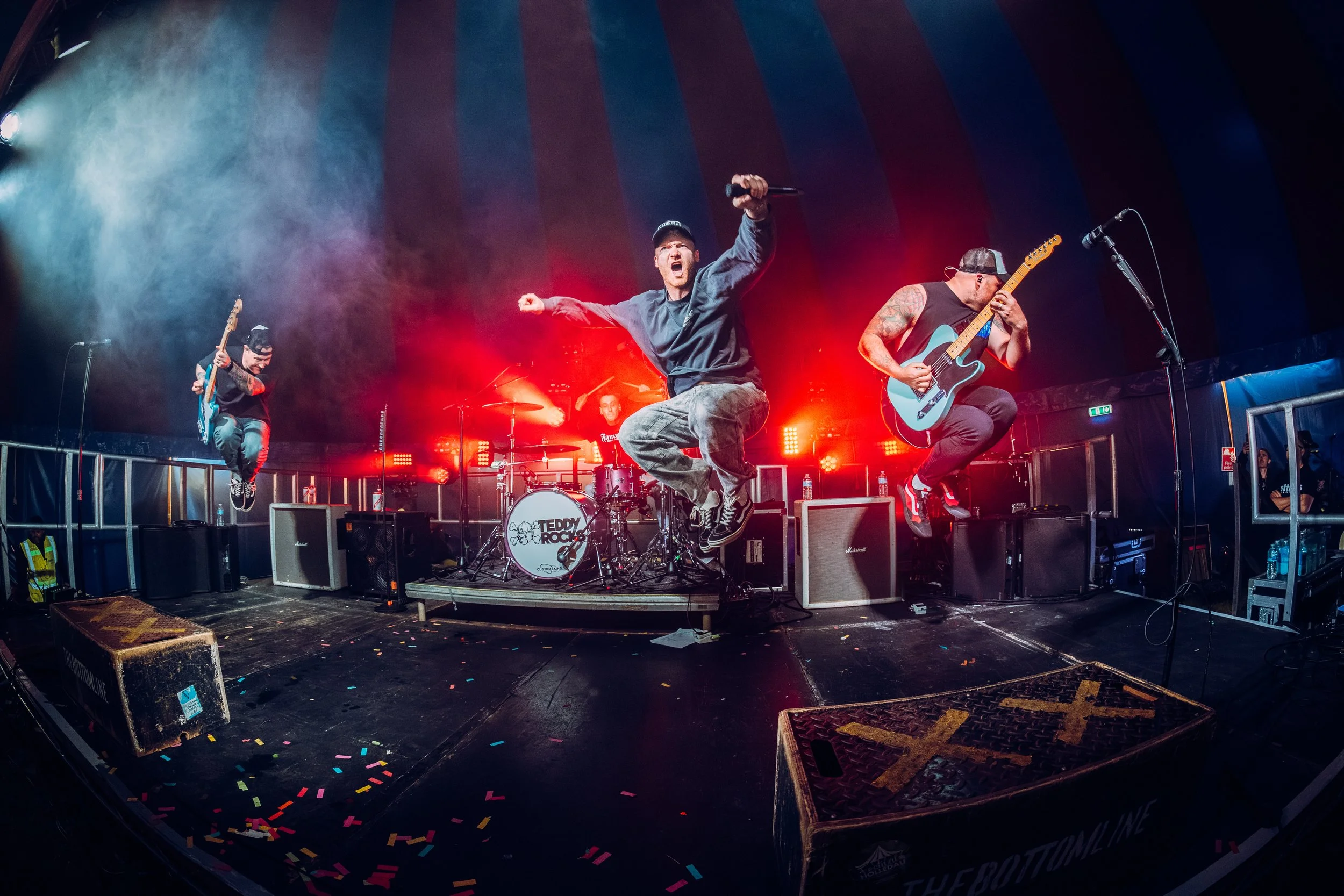 A dynamic wide fisheye perspective capturing pop-punk band The Bottom Line executing synchronized mid-air stage jumps under a festival tent at Teddy Rocks Festival. High-action music festival stage performance and entertainment photography by Will Ba