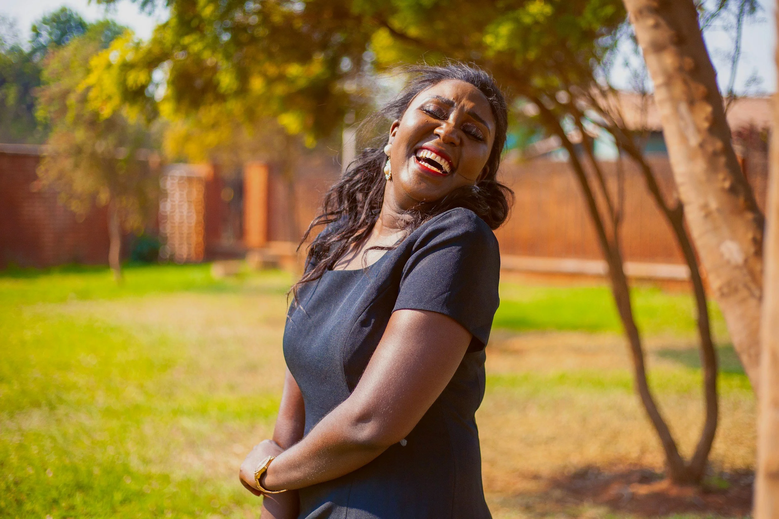 A woman with dark hair and makeup smiling and laughing outdoors on a sunny day, standing near a tree with a brick background and lush green grass.