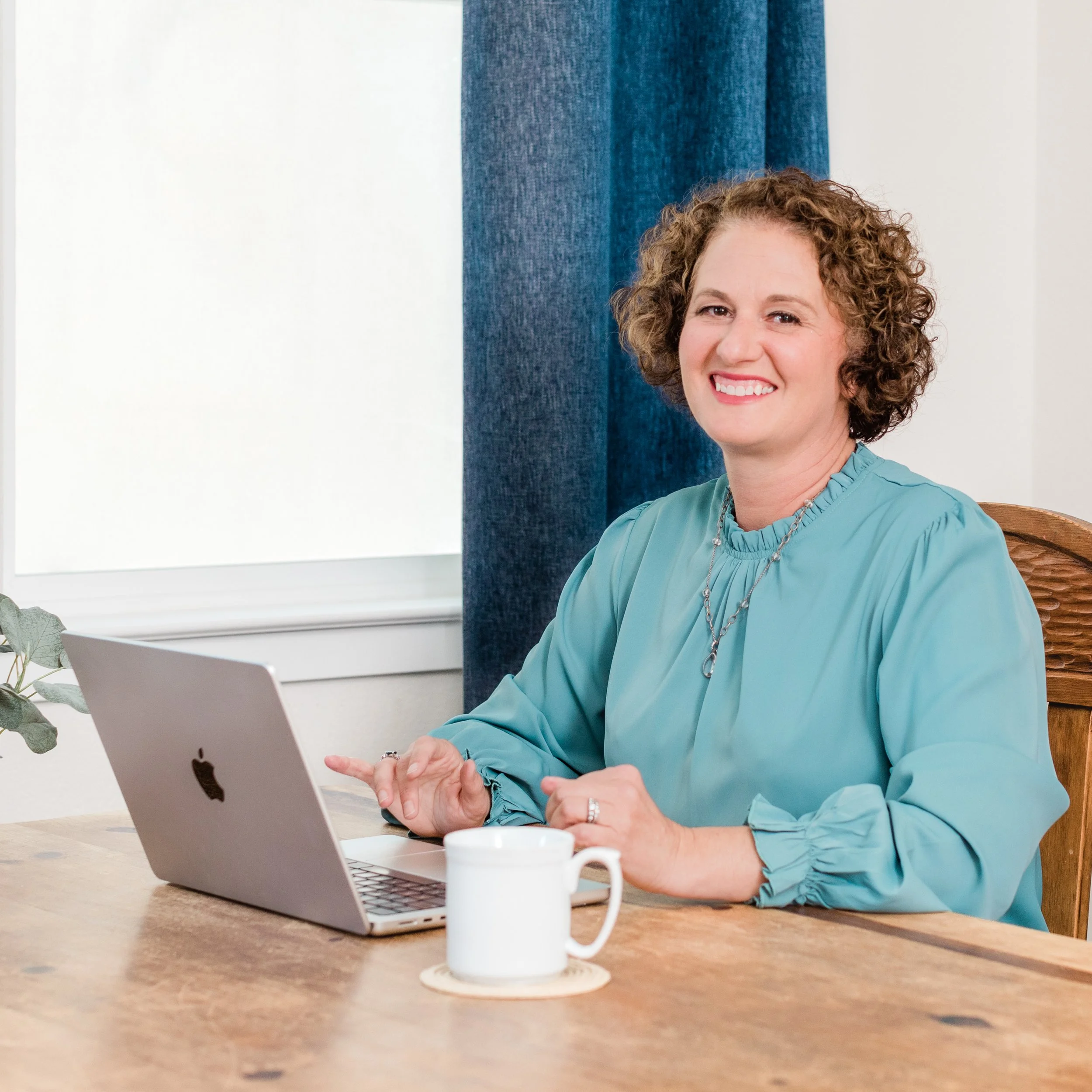 Devon Vernetti with curly hair and a blue blouse smiling while sitting at a wooden table with an open laptop. There is a white mug in front of her and a blue curtain behind her.