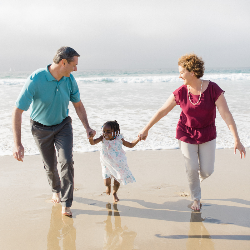 Family of three, a man, woman, and young girl, holding hands and playing on the beach near the shoreline with waves in the background, sunny weather.