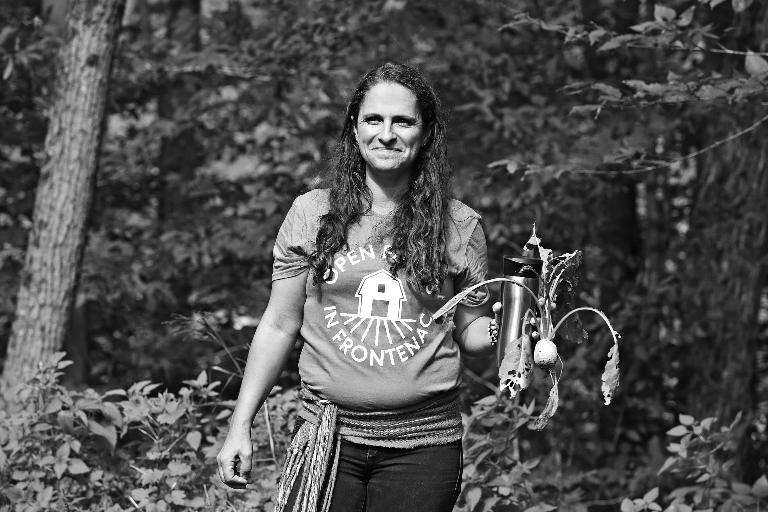 A woman with long hair holding a plant with a vegetable and a thermos, standing outdoors in a forested area.