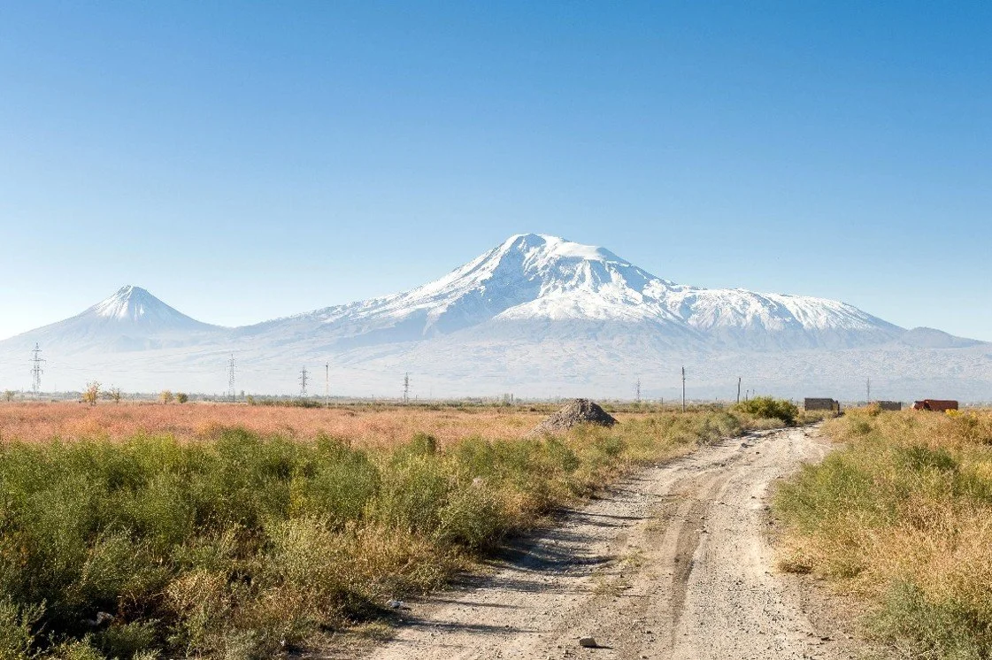 Snow-capped Mount Hood and Mount Adams in the distance, viewed over a dirt road through a grassy field.