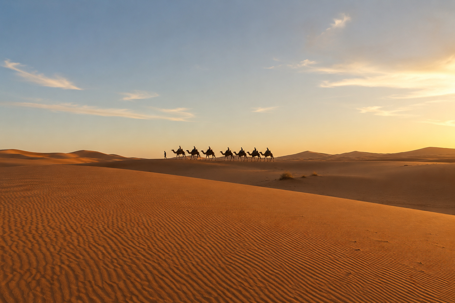 A desert landscape at sunset with sand dunes and a caravan of seven camels with riders, with a person leading the camels.