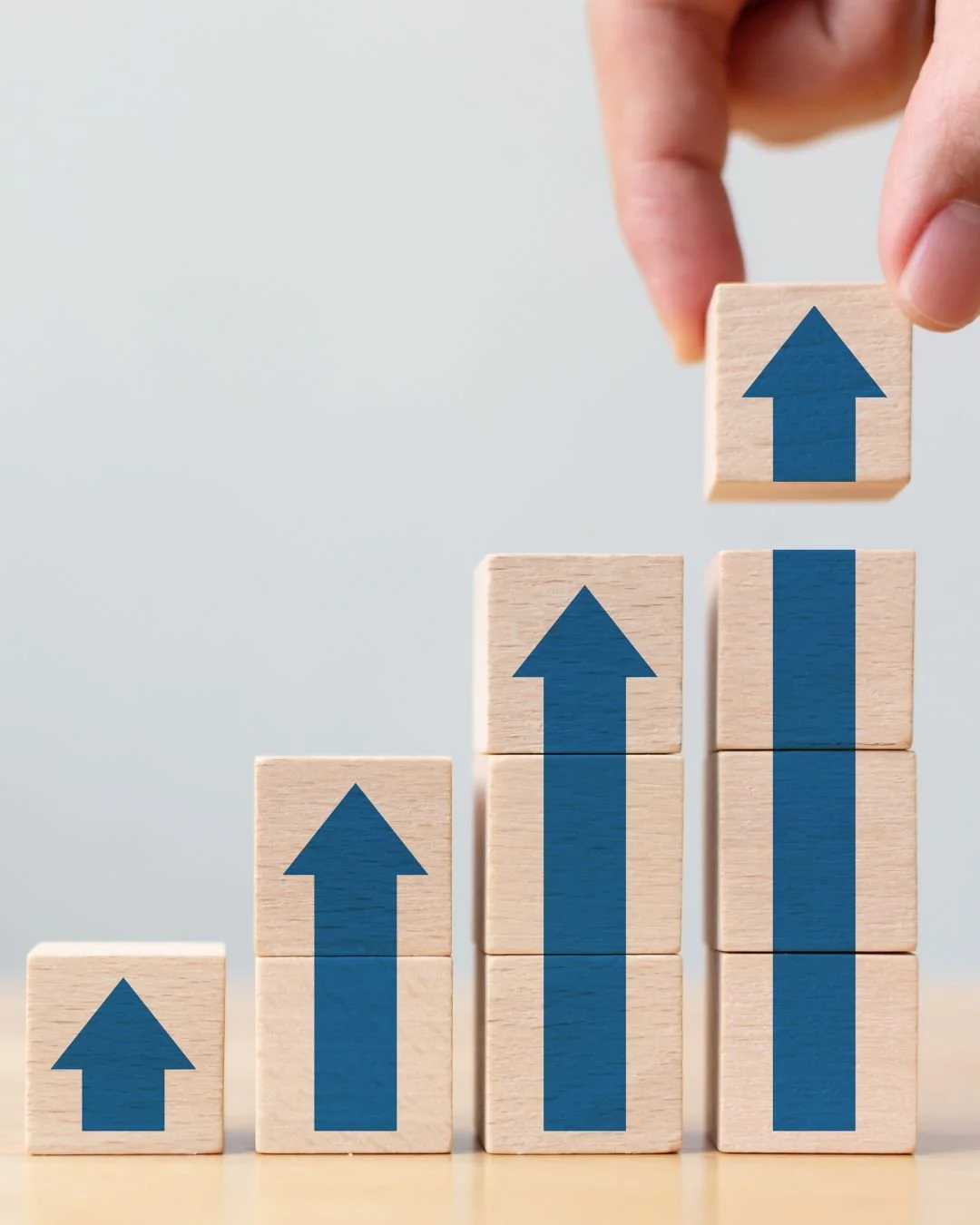 Wooden blocks with blue upward arrows, arranged in increasing height from left to right, with a hand placing the tallest block.