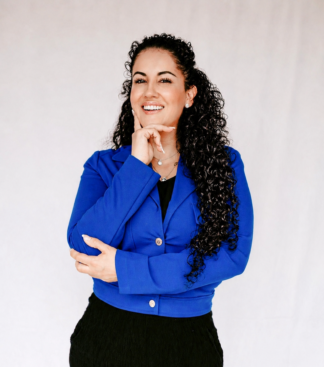 A woman with long, curly black hair wearing a blue jacket and black top, smiling with her hand on her chin, standing against a plain white background.