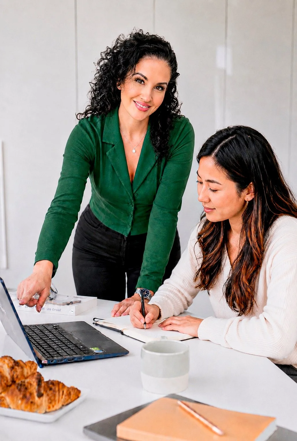 Two women in an office setting, one standing and pointing at a laptop screen, the other seated and writing in a notebook, with a plate of croissants on the table.