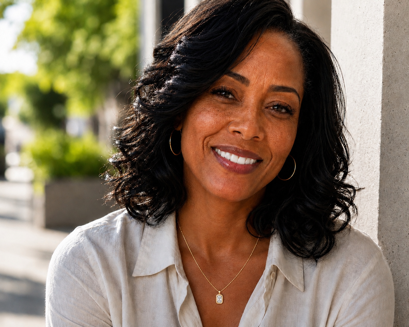 A smiling woman with black curly hair, wearing hoop earrings and a gold necklace, standing outdoors next to a wall with greenery in the background.