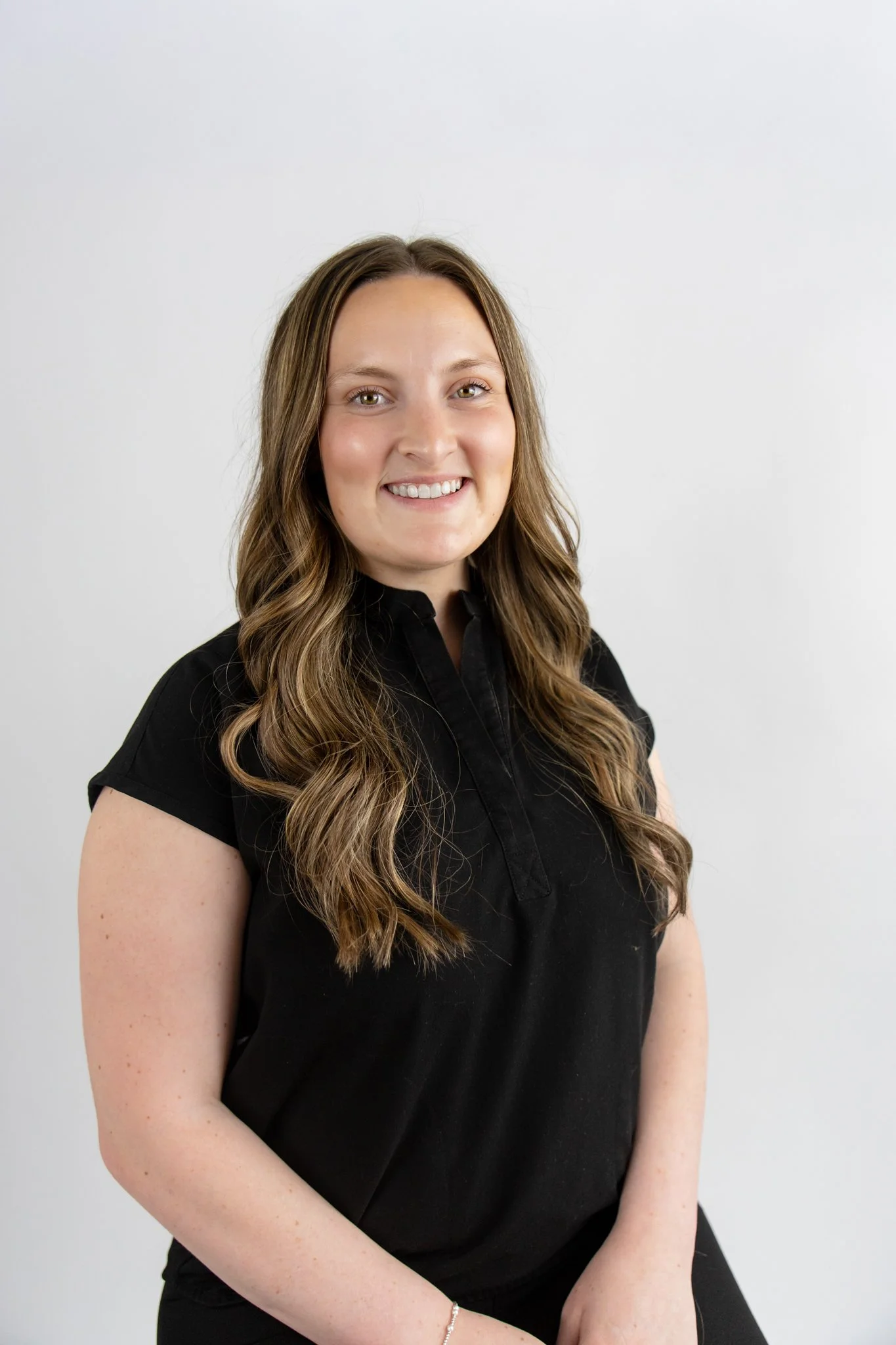 A woman with long wavy brown hair with blonde highlights wearing a black shirt, smiling, against a plain white background.