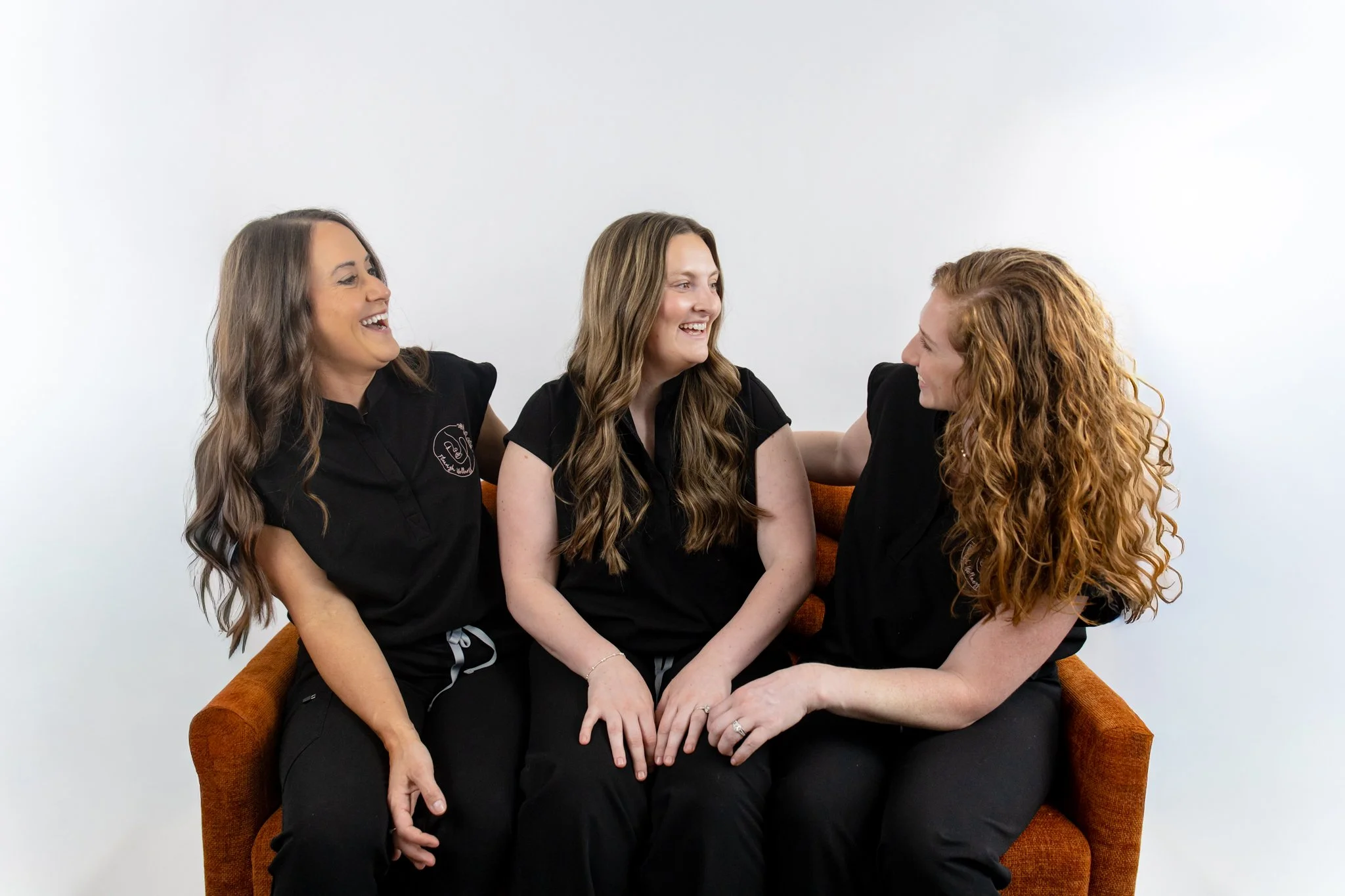 Four women with long hair, two with brown and two with red, sitting close together on an orange couch, smiling and engaging in conversation against a plain white wall background.