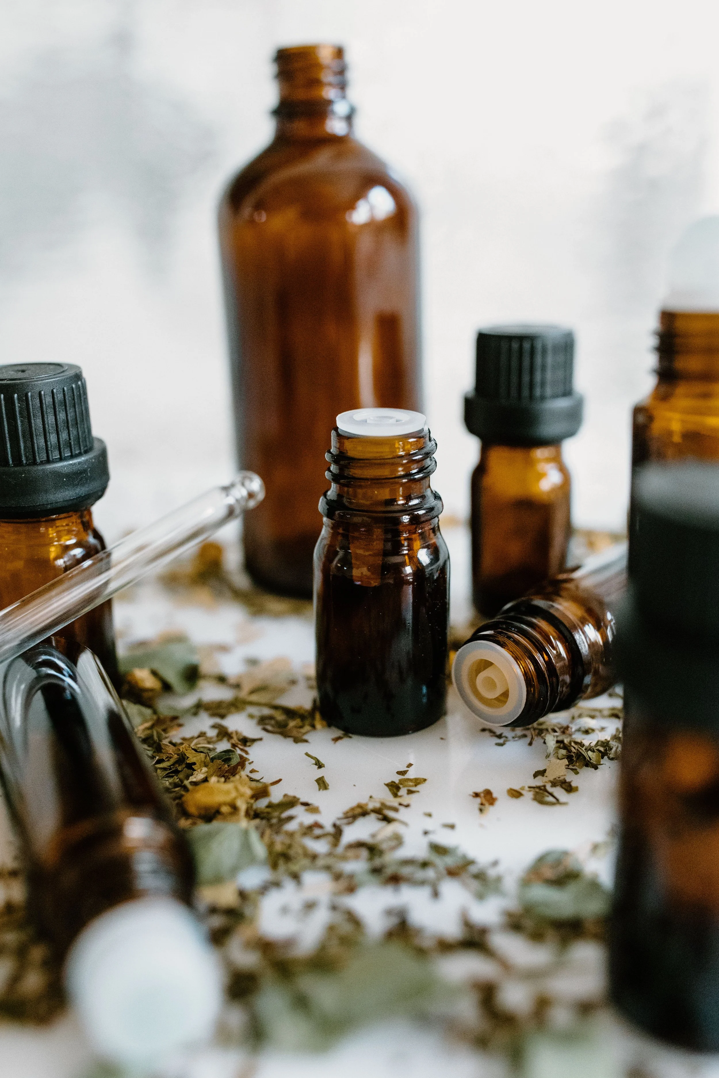 Various small brown glass bottles and jars with black caps, some lying down, surrounded by dried herbs or tea leaves on a white surface.