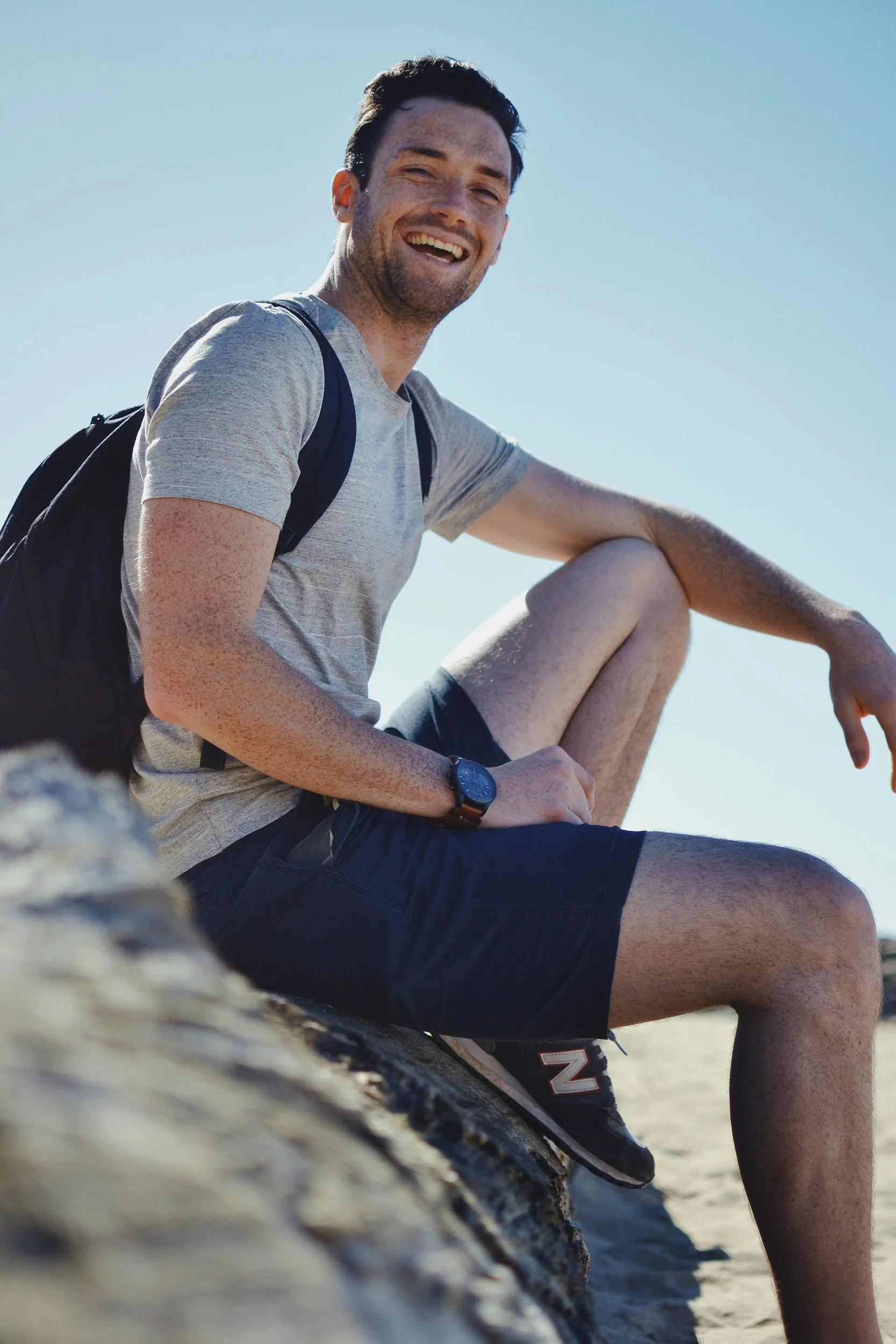 Young man smiling while sitting on rocks at the beach, wearing a gray t-shirt, black shorts, and a black backpack, with clear blue sky in the background.