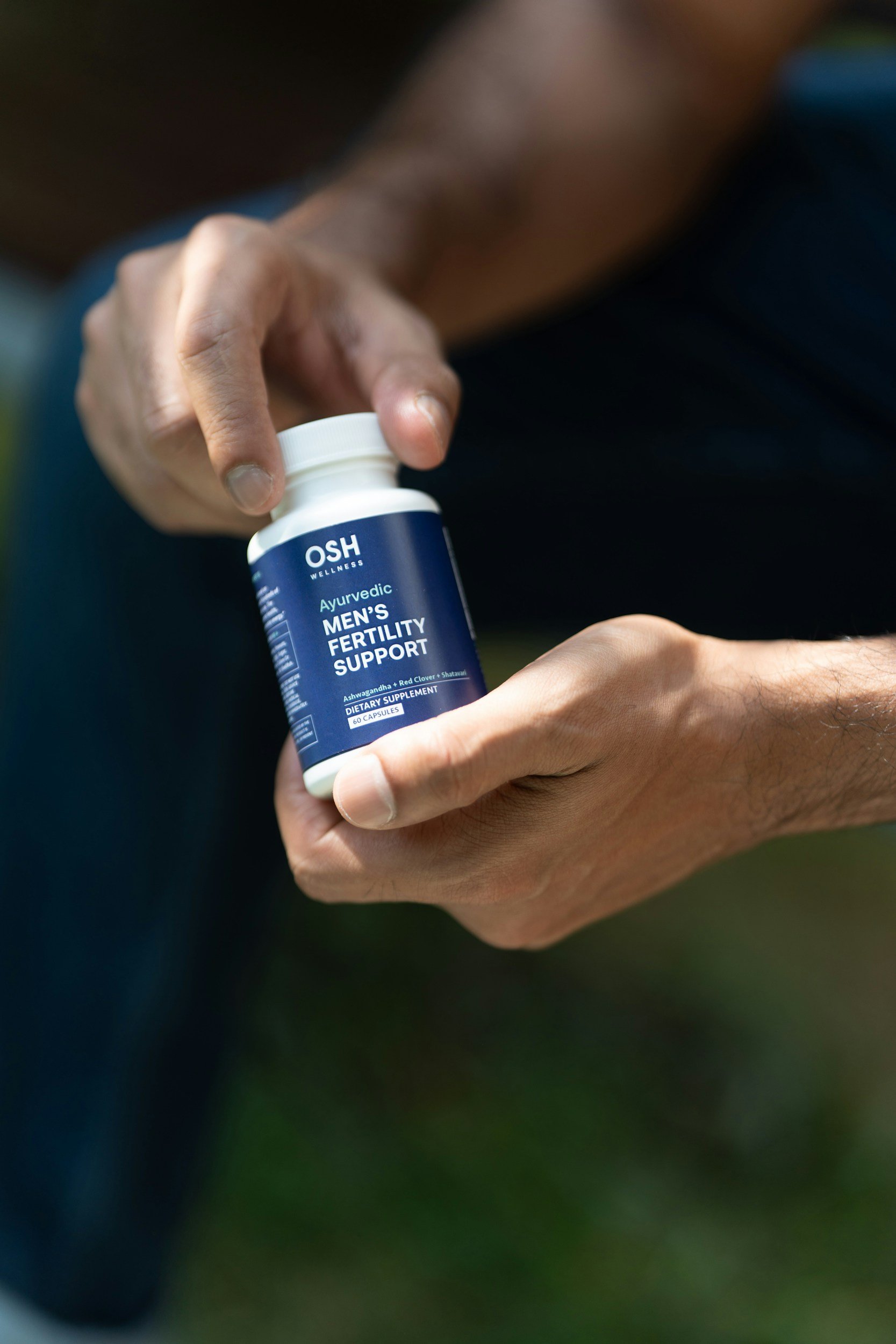 Close-up of a person holding a bottle of OSH Wellness Ayurvedic men's fertility support dietary supplement capsules outdoors.